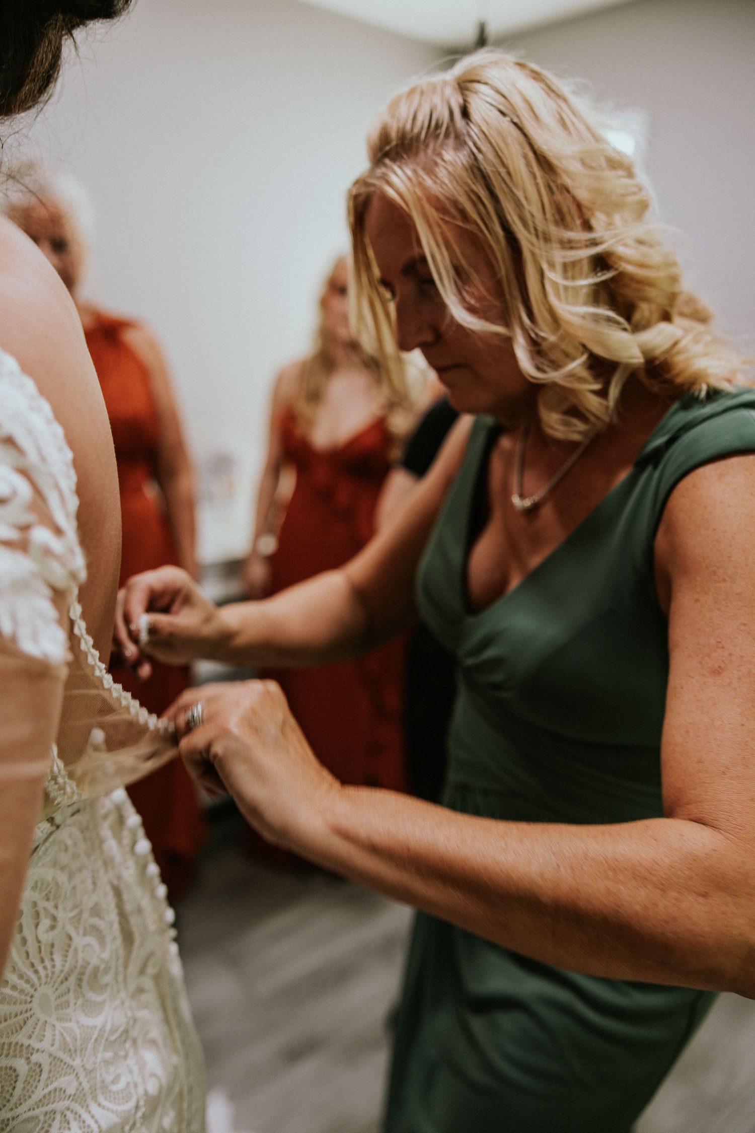 Blonde woman in green dress buttoning the back of a lace wedding gown while bridesmaids watch in background.