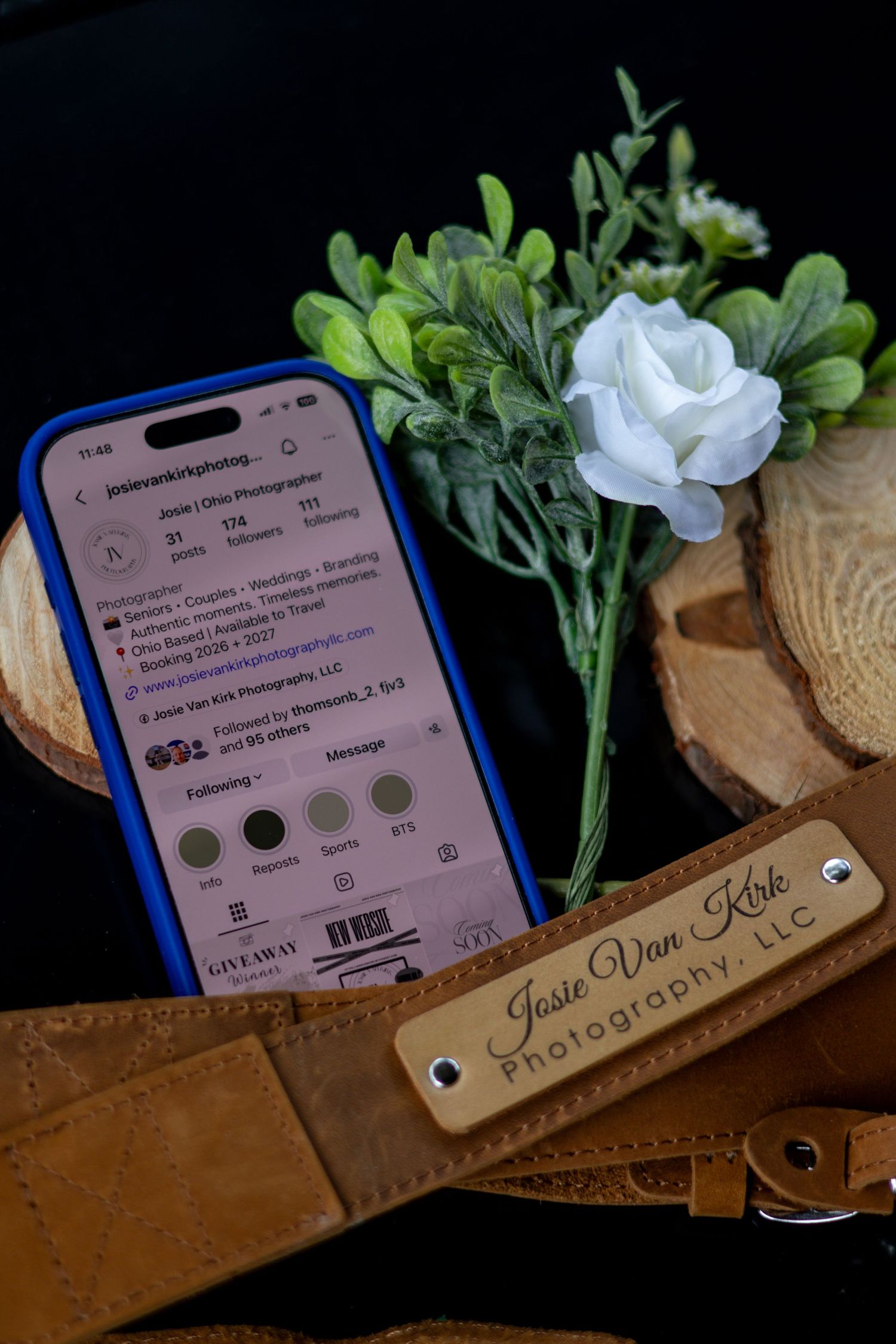 Jessie Van Kirk Photography LLC Instagram profile on phone beside white rose and branded leather strap.