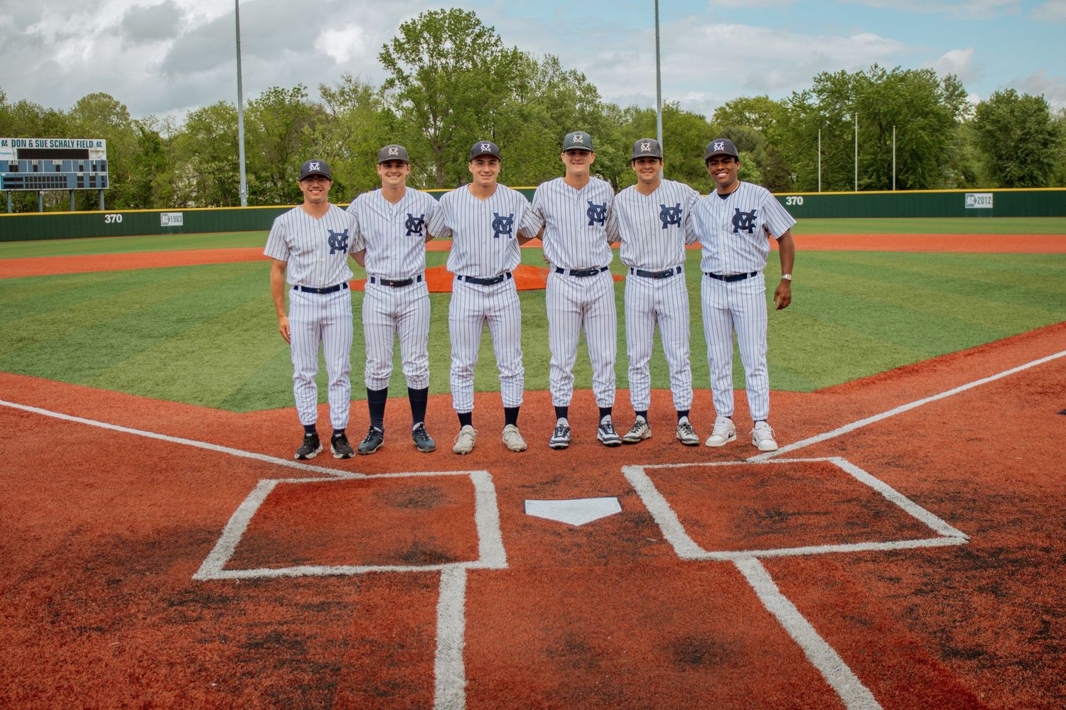 Six baseball players in white uniforms standing at home plate on a baseball diamond.