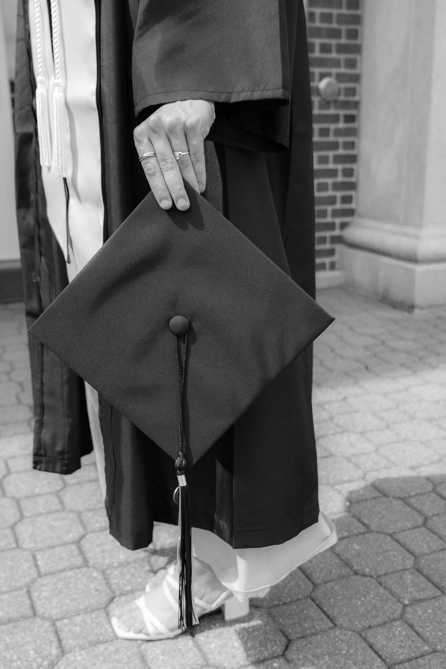 Black and white photo of graduate in cap and gown holding mortarboard by brick building.