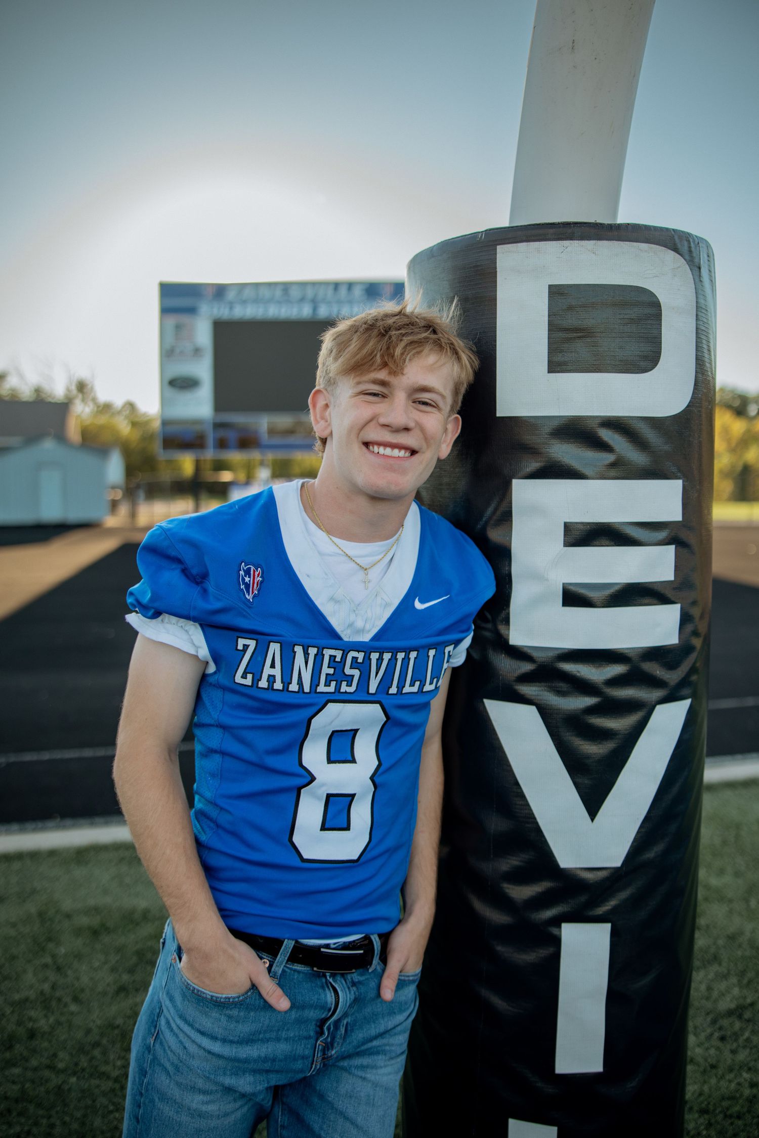 Smiling teen in Zanesville #8 blue football jersey poses next to a Devils sign on a football field.