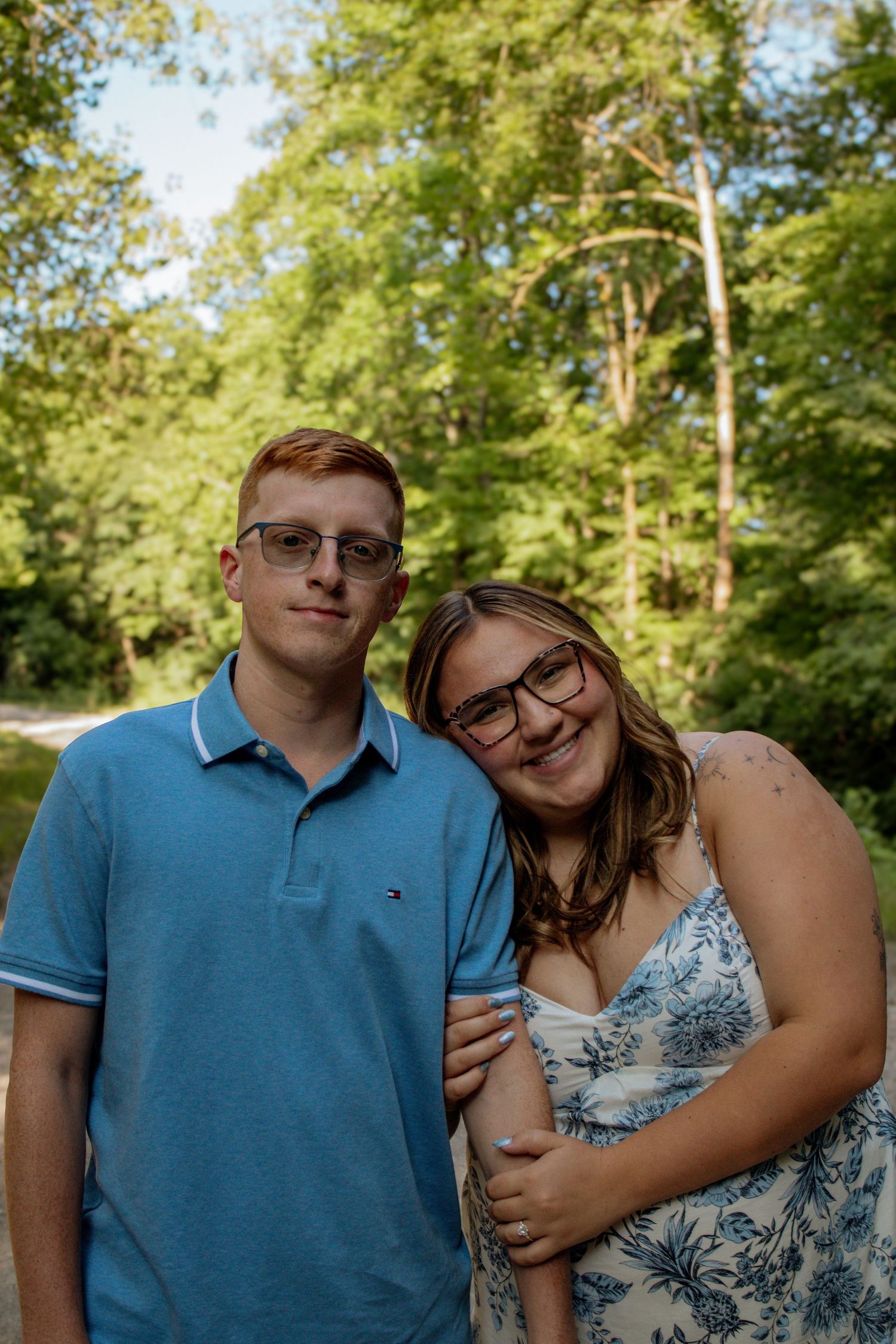 Young couple with glasses smiling together outdoors surrounded by lush green trees on sunny day.