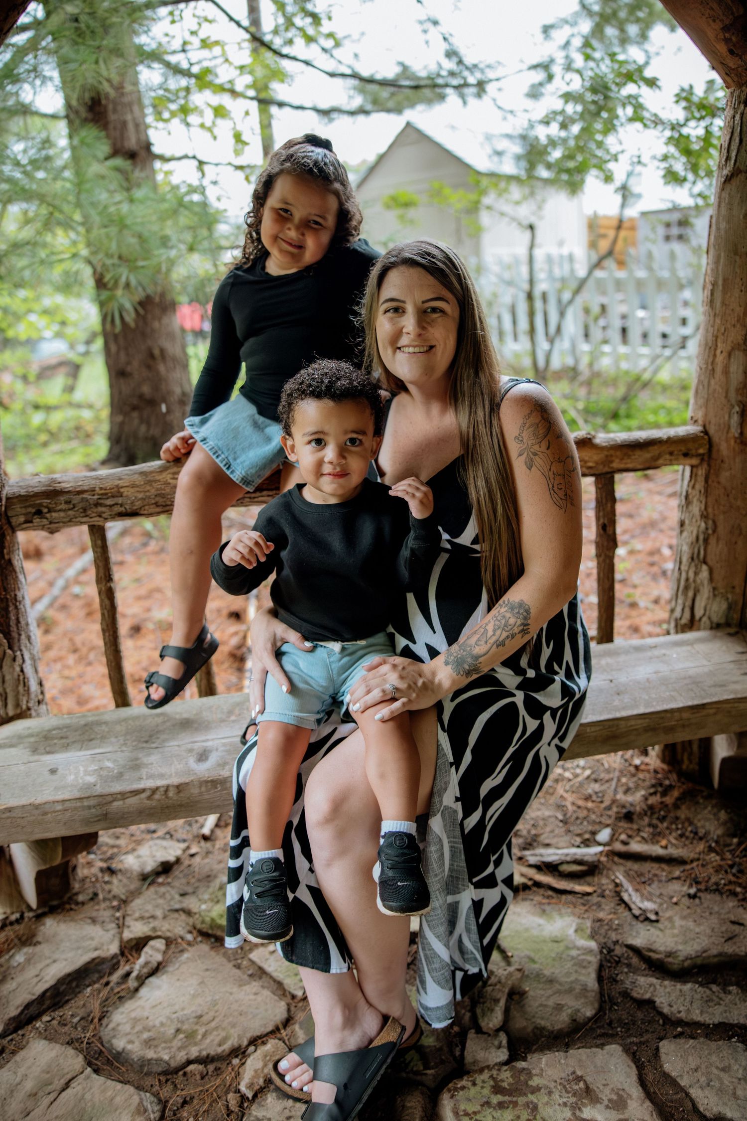 A woman sits outdoors on a wooden bench with two young children on her lap, smiling in a lush garden setting.