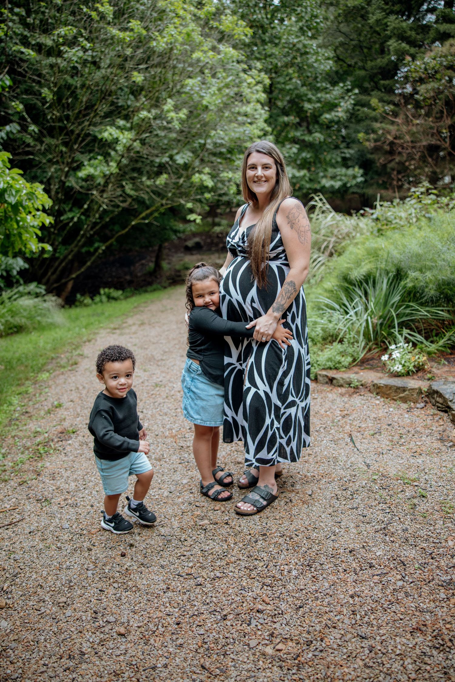 Pregnant woman in black and white dress poses with two young children on a garden path surrounded by lush greenery.
