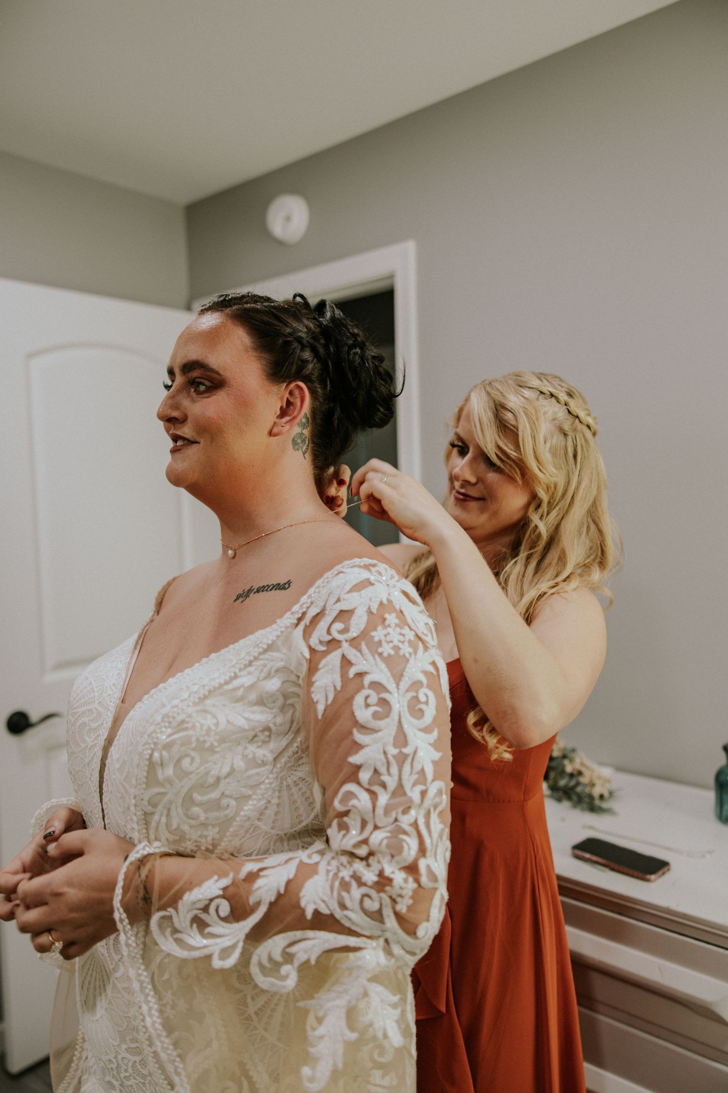 Bridesmaid in rust dress helping bride with earring as she wears lace long-sleeve wedding gown.