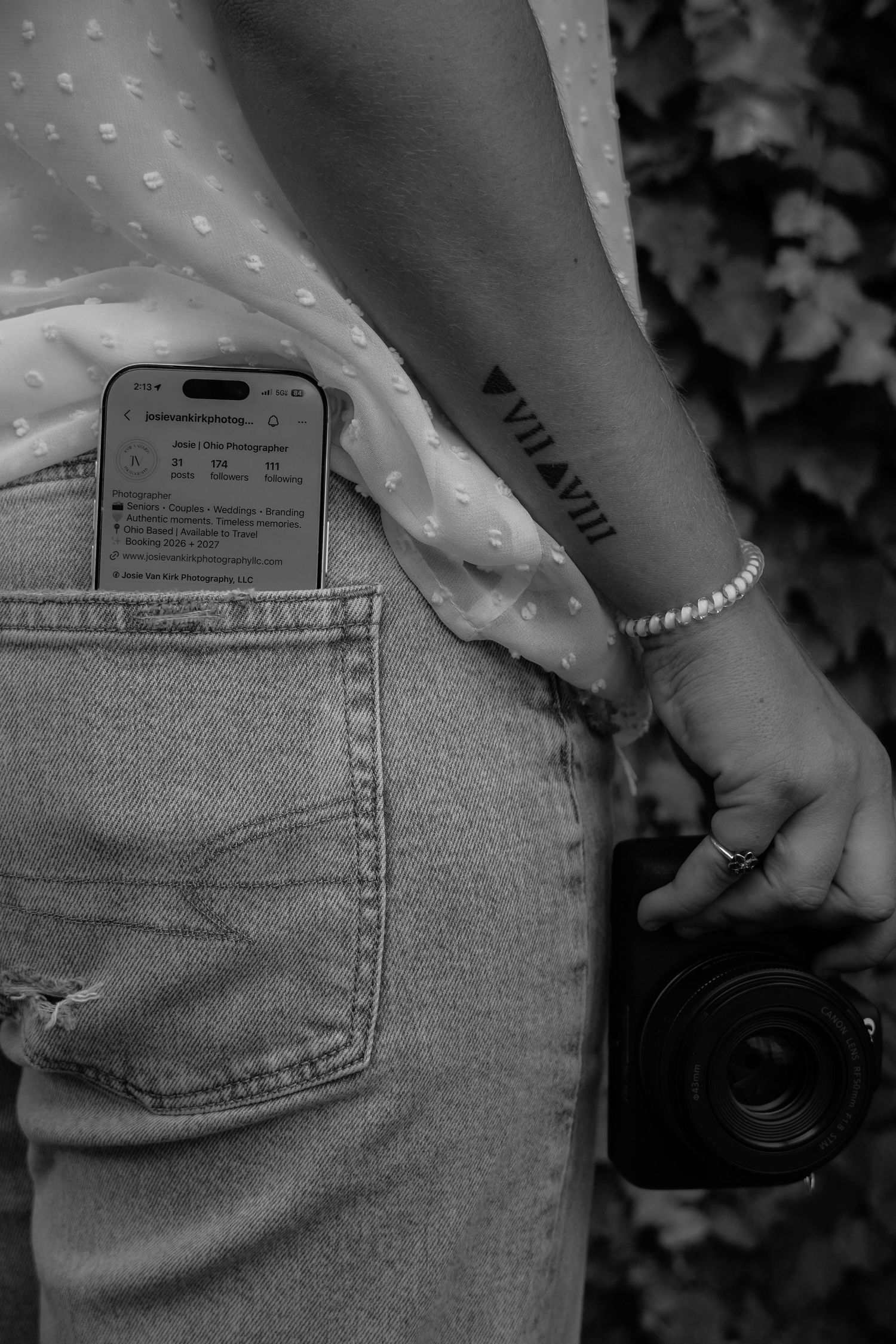 Black and white photo of photographer holding camera with phone in back pocket of ripped jeans.