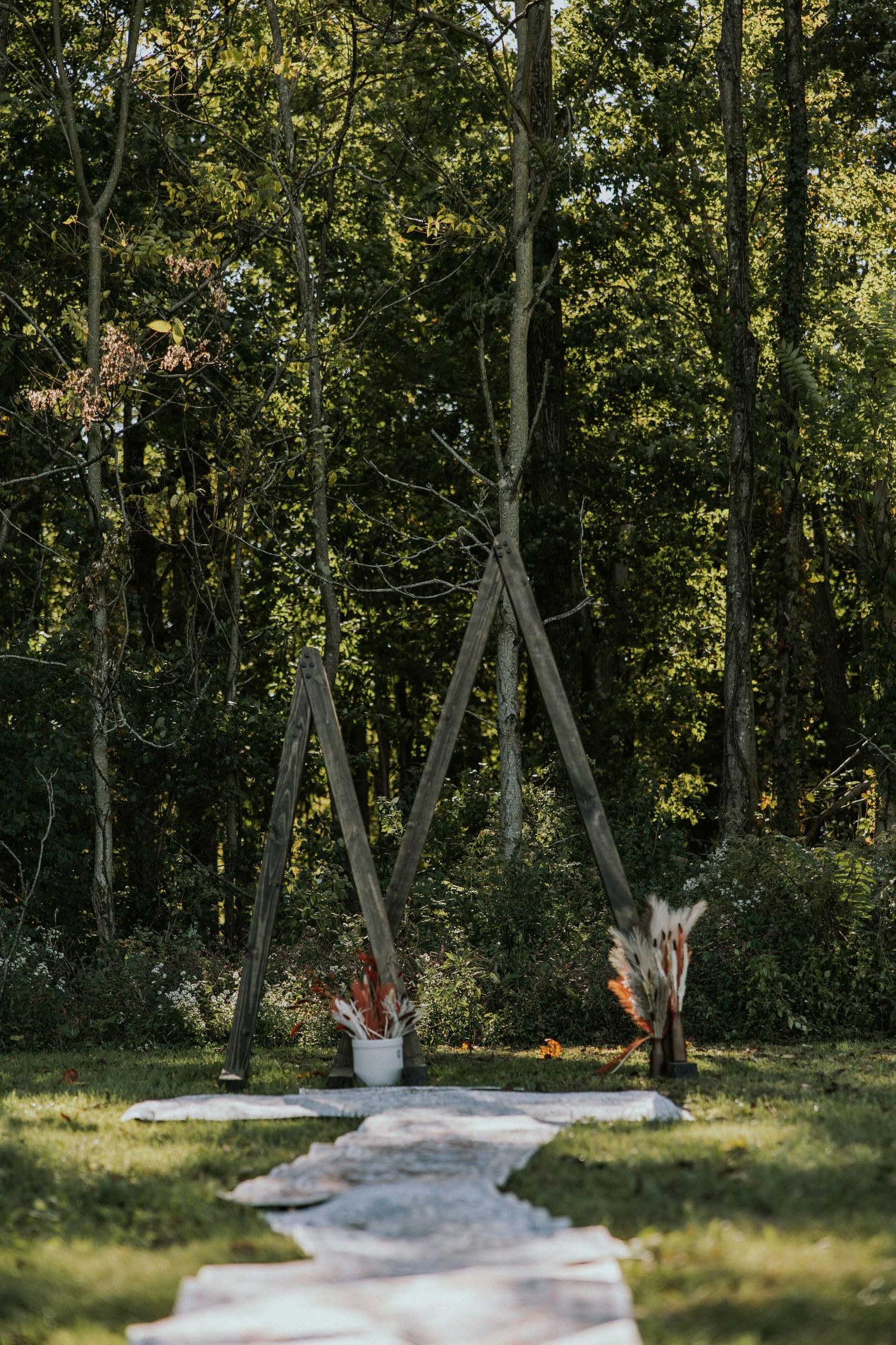 Rustic wooden triangular wedding arch with pampas grass arrangements on a stone path surrounded by lush green trees.