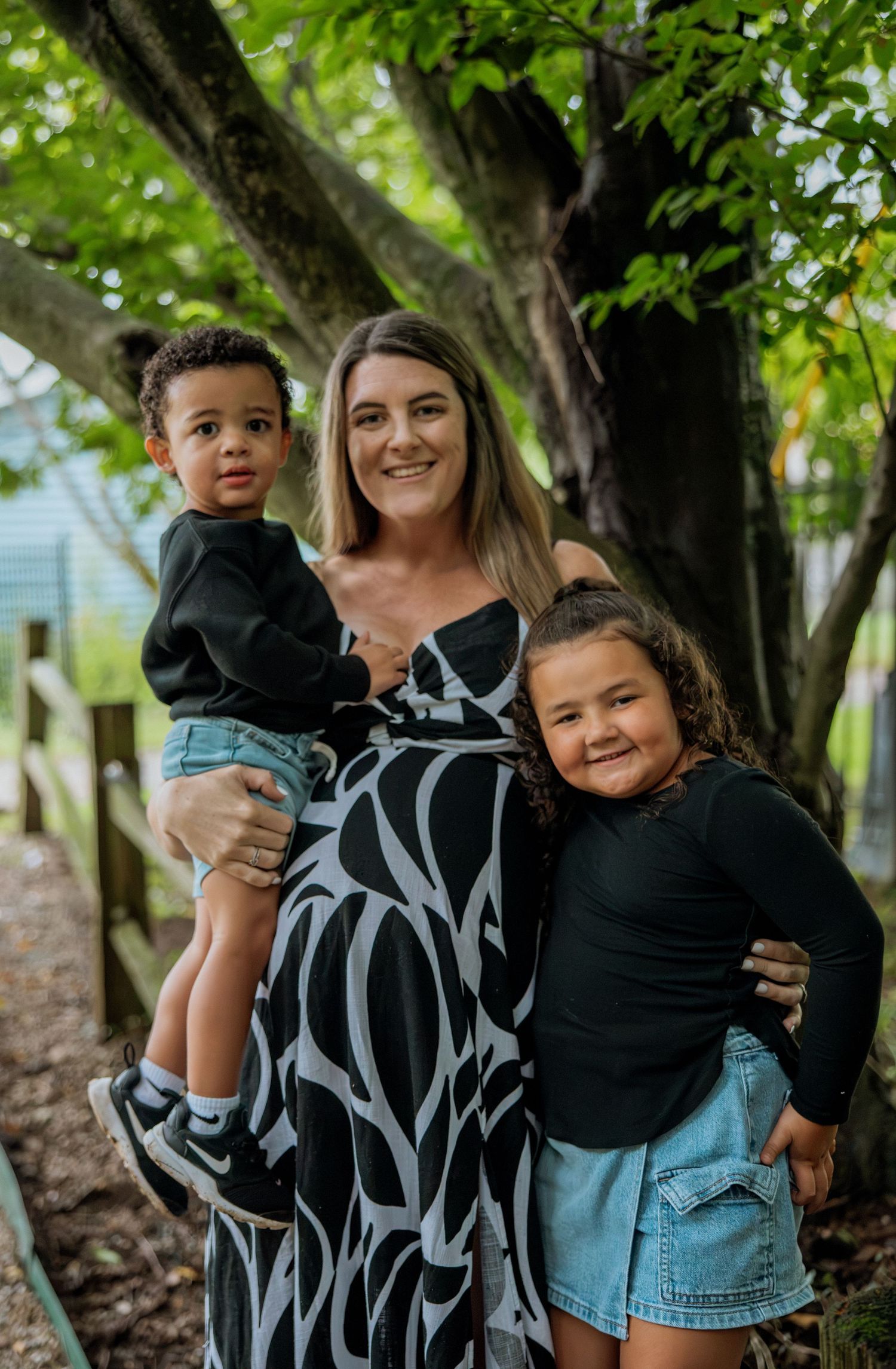 A smiling woman in a black and white dress holds a toddler boy while a young girl in black hugs her side outdoors.