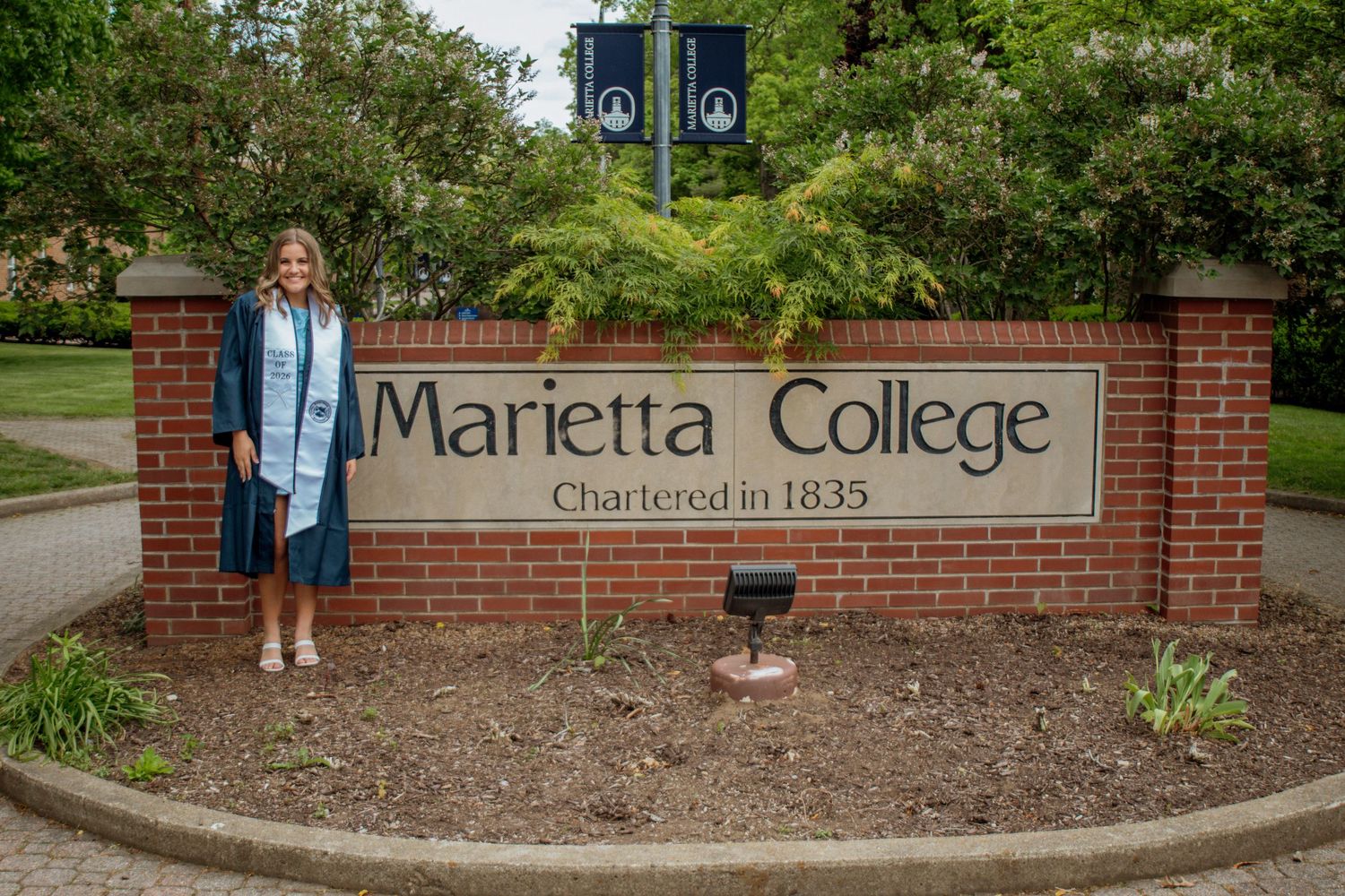 Smiling graduate standing beside Marietta College chartered 1835 brick sign.