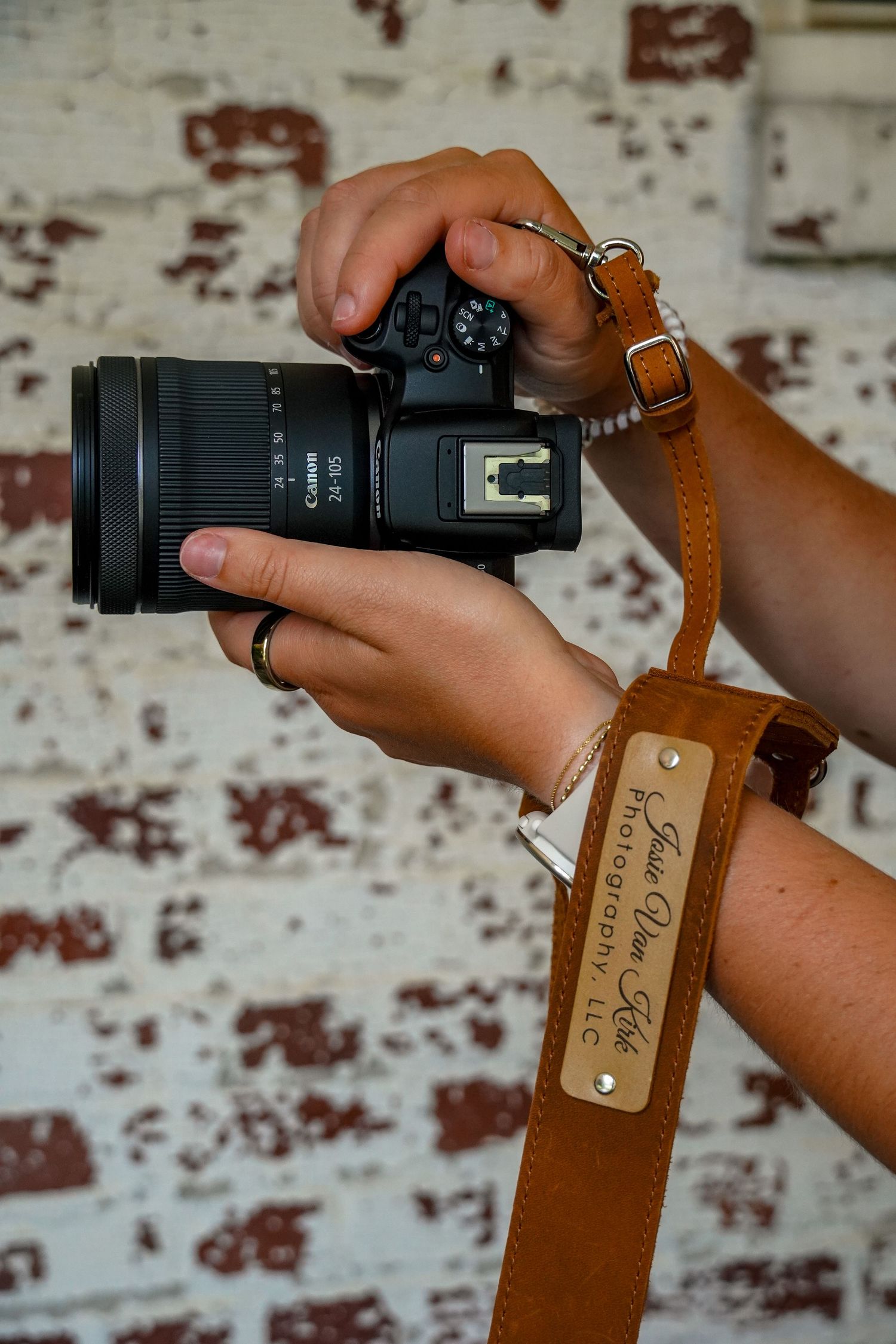 Hands holding Canon camera with tan leather Jessie Van Kirk Photography LLC strap against brick wall.