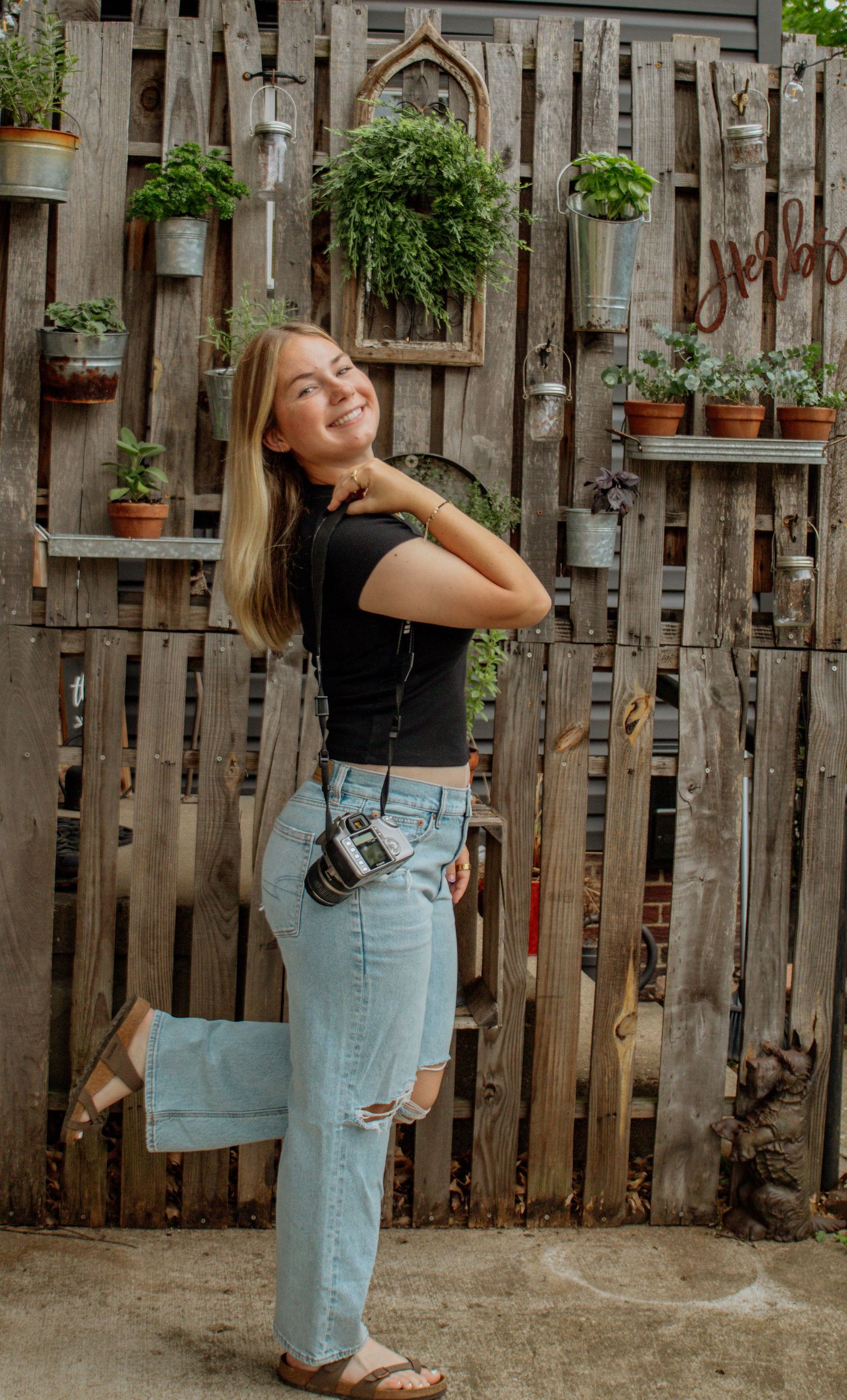 Smiling blonde female photographer posing with Canon camera in front of rustic wooden wall with hanging potted plants.