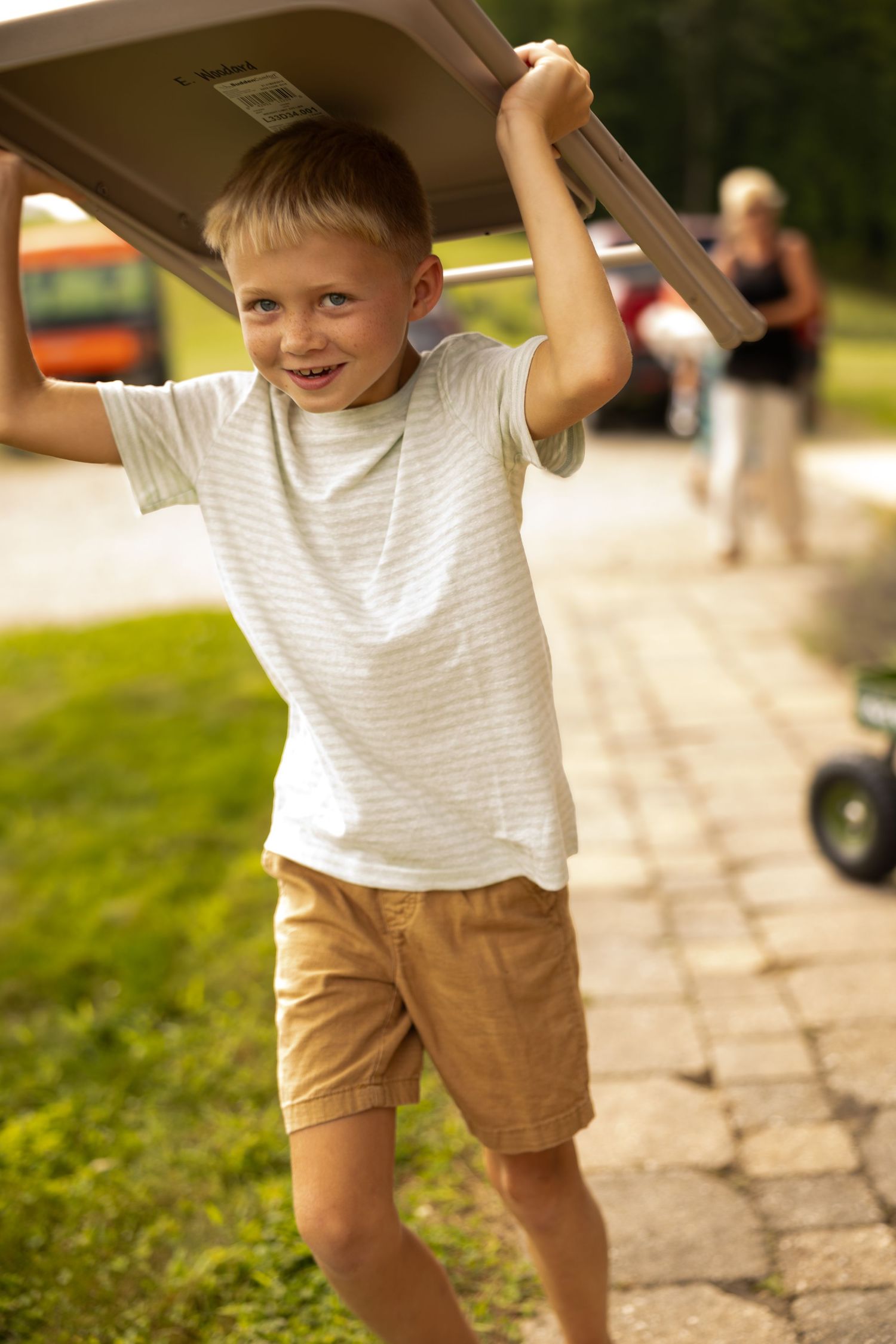Happy young boy carrying a folding table overhead on a sunny day outdoors, wearing a striped shirt and tan shorts.
