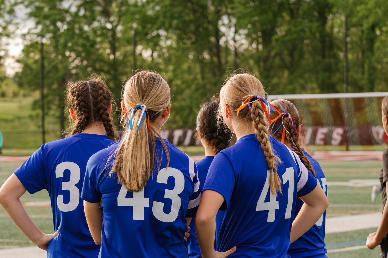 Four girls in blue soccer jerseys with numbers 3, 43, and 41 stand together on a sports field.