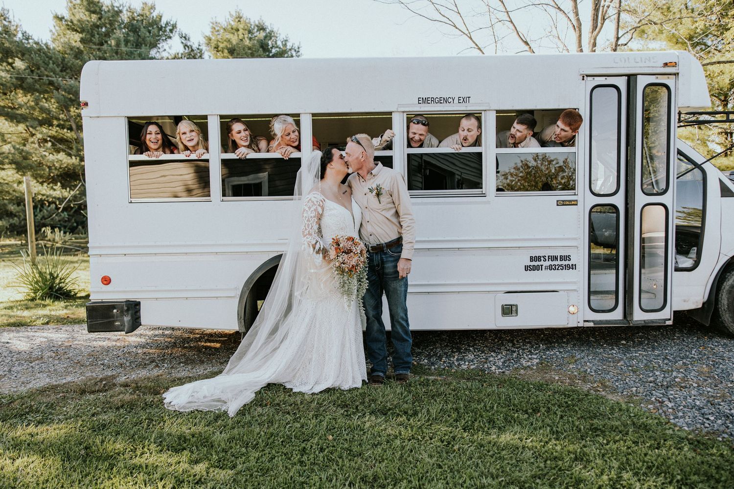Bride and groom kissing in front of a white vintage party bus with wedding guests peering through windows.
