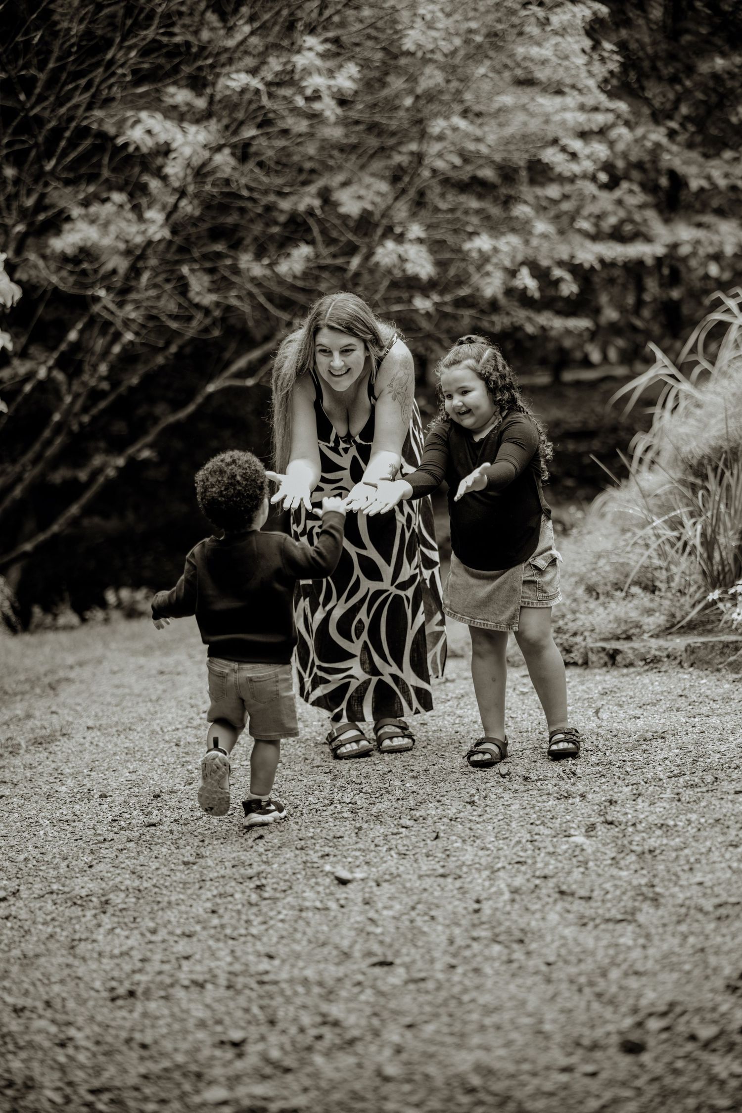 Black and white photo of a smiling woman bending down to greet two young children on a gravel path outdoors.