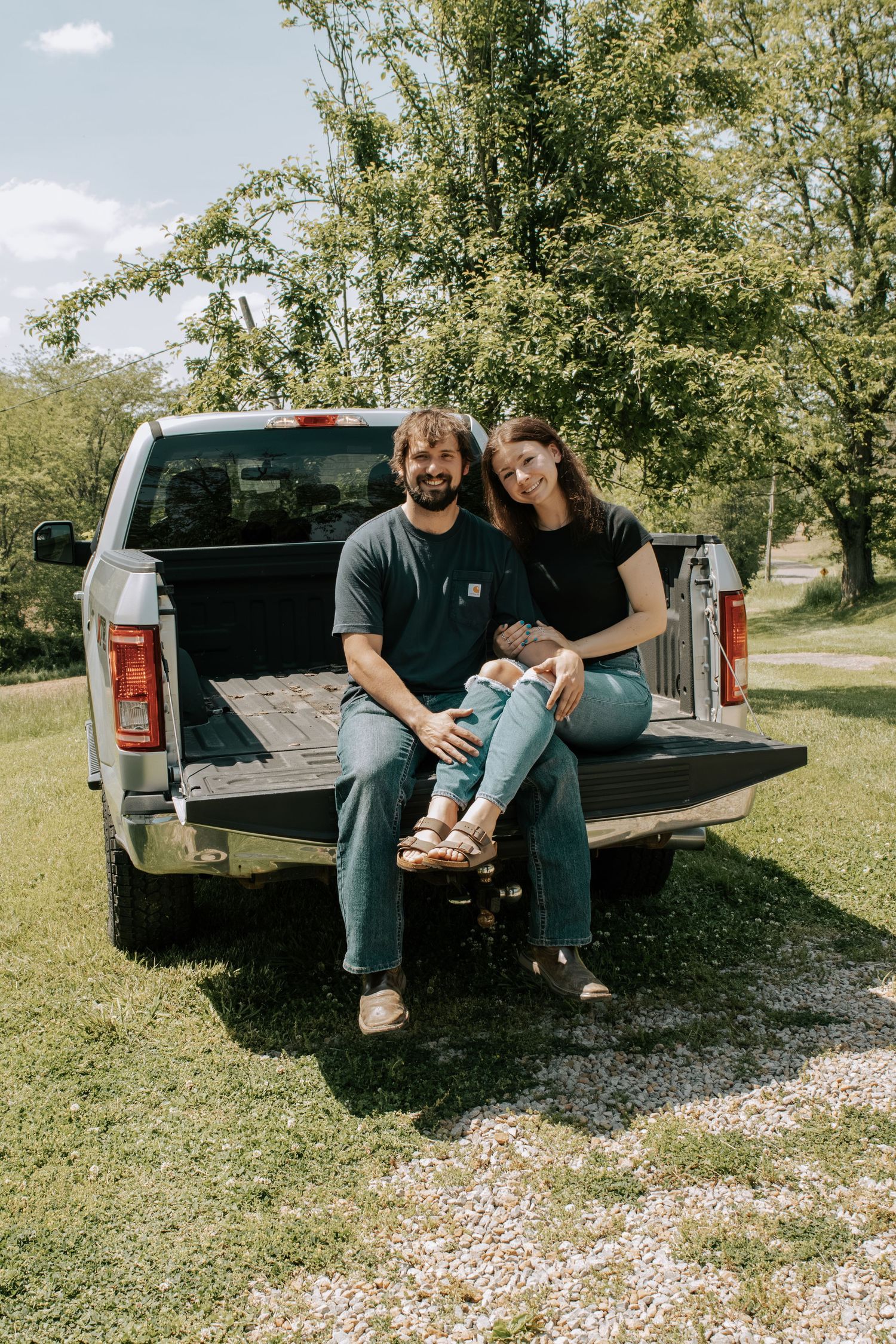 A happy couple sits together on a pickup truck tailgate in a sunny green yard.