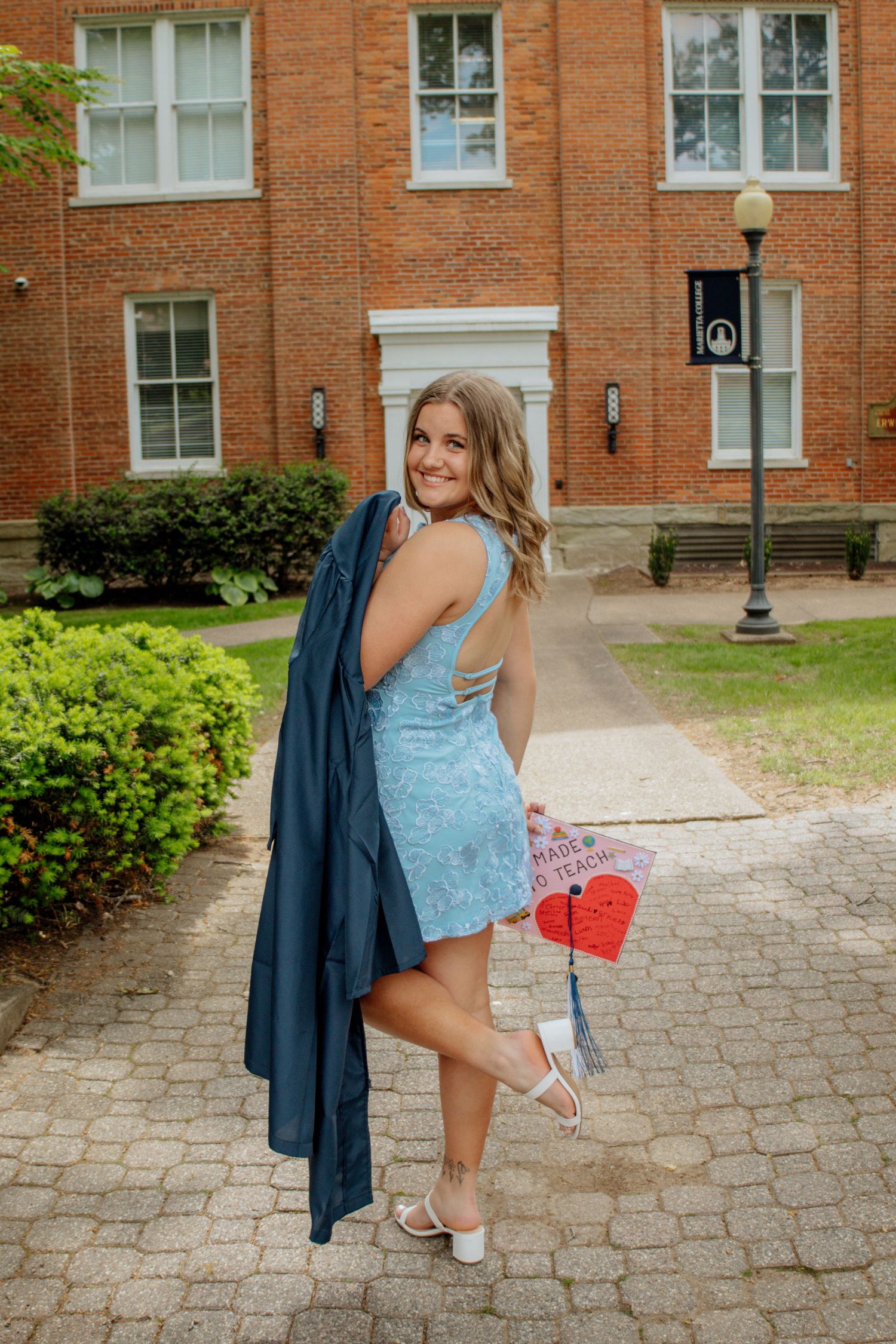 Smiling female graduate in blue dress holds navy gown over shoulder on brick campus path.