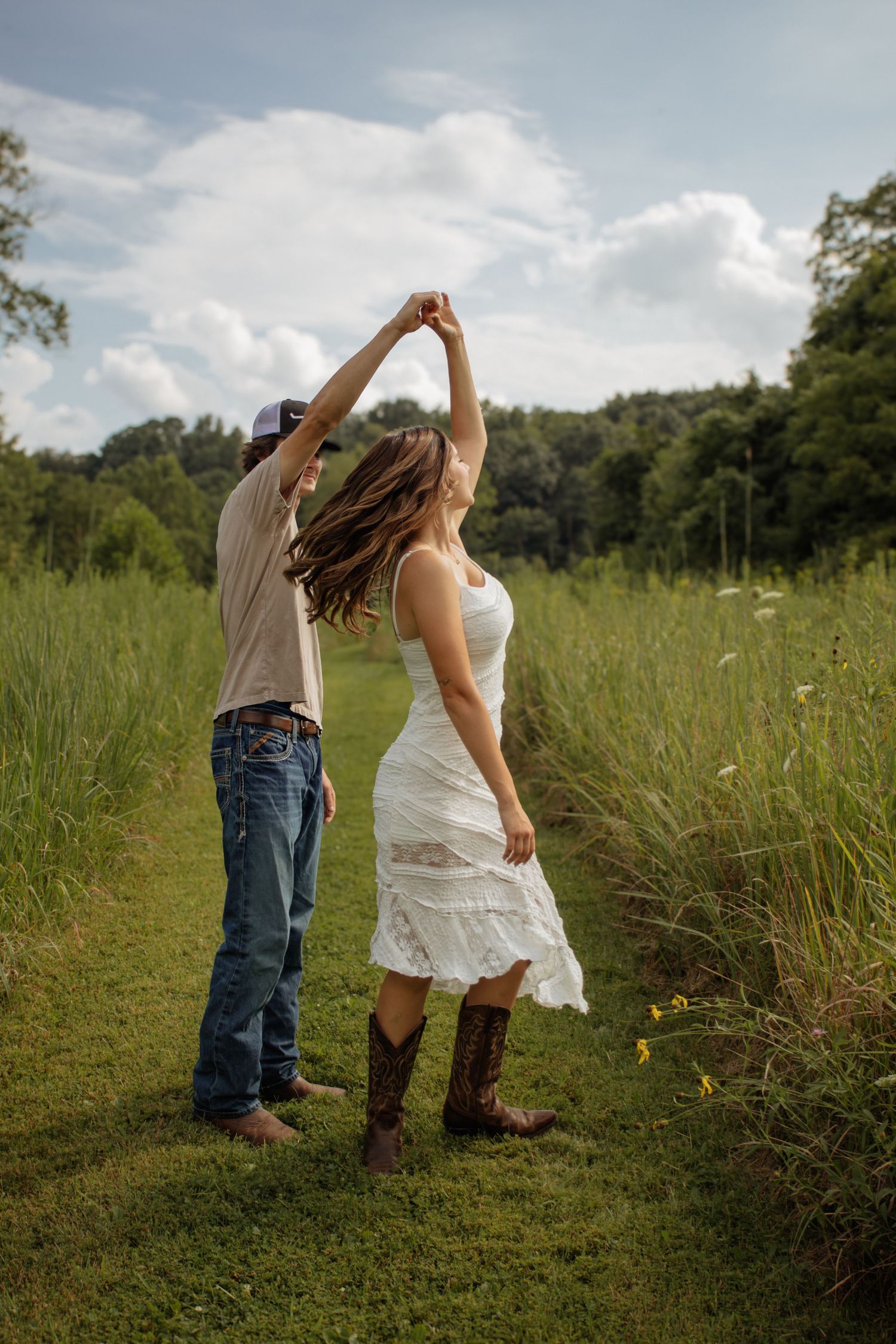 Couple dancing outdoors in a green field, man in jeans and cap spinning woman in white lace dress and cowboy boots.