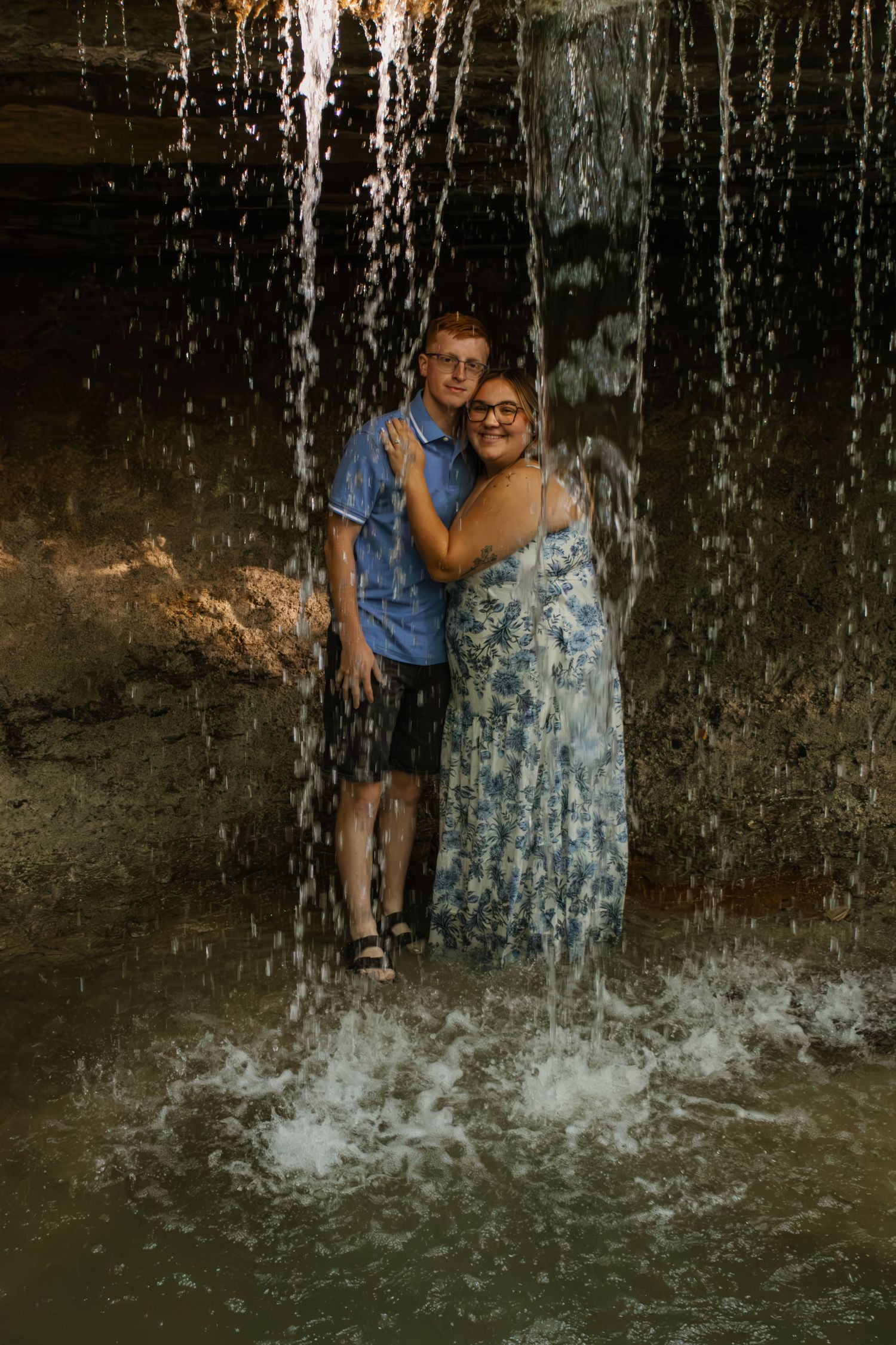 A smiling couple embraces behind a curtain of falling waterfall water in warm light.