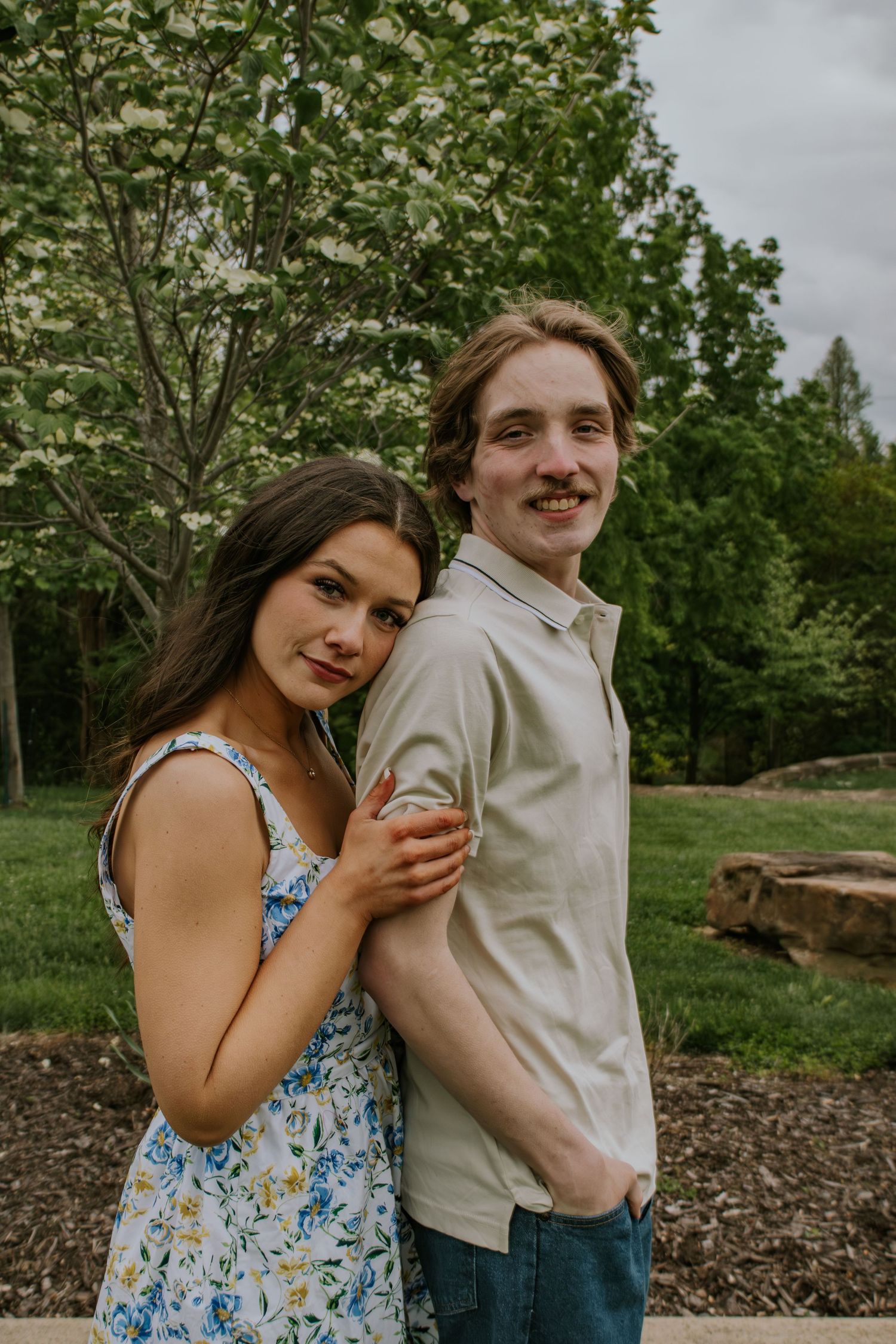 Smiling couple posing back-to-back in a park with blooming white trees and green foliage.