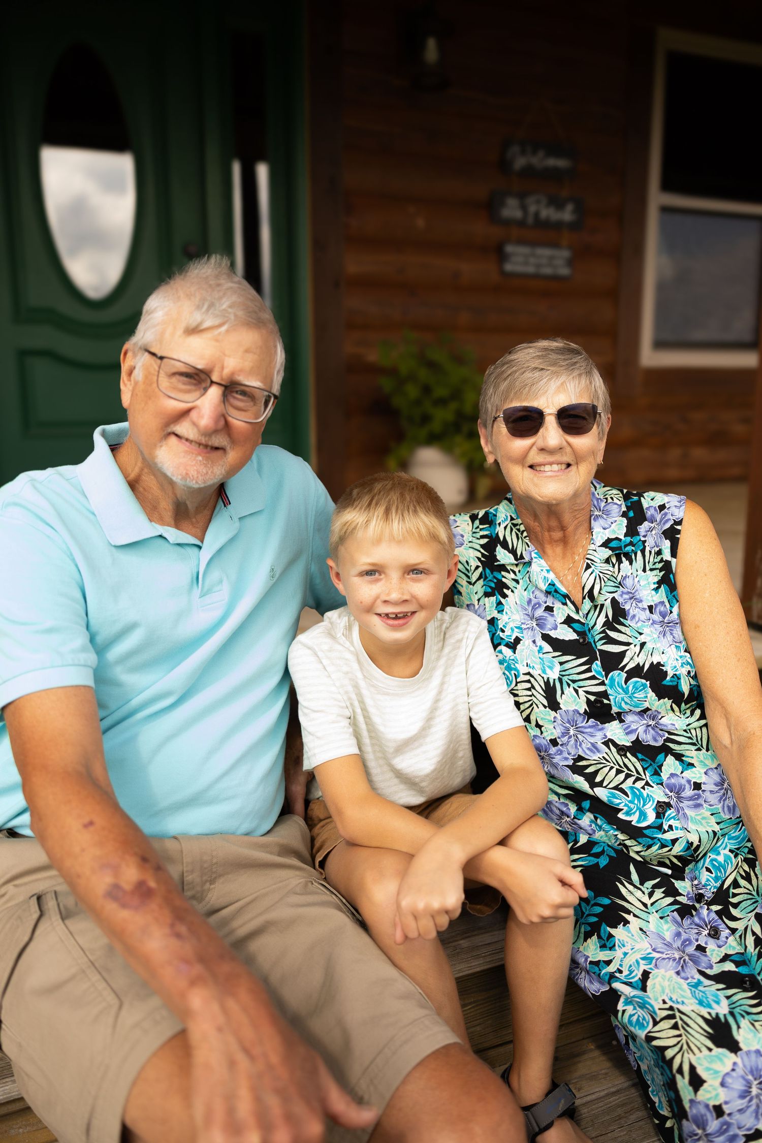 Elderly grandparents sitting on porch steps with young grandson, smiling outdoors near green front door.