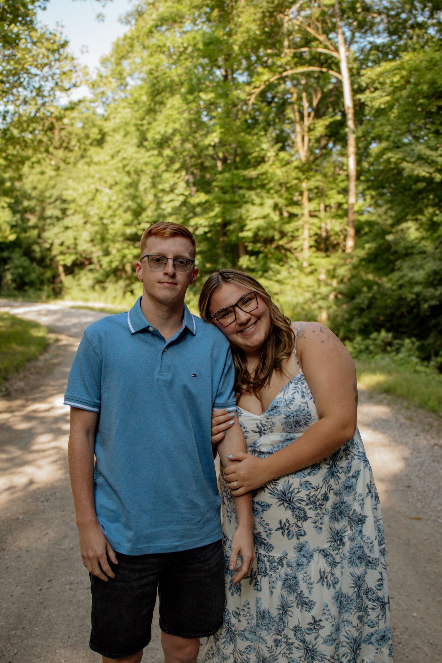 A young couple poses together on a scenic wooded gravel path on a sunny day.
