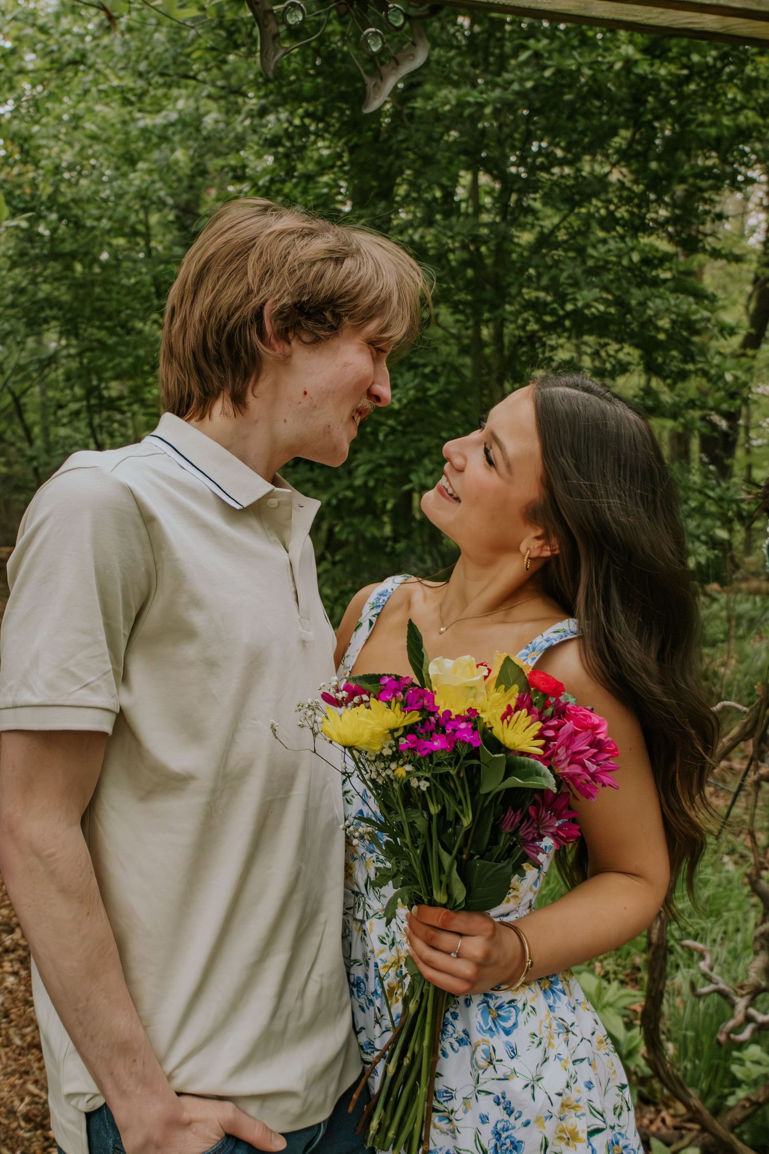 Young couple smiling at each other as woman holds colorful bouquet of flowers in a garden setting.