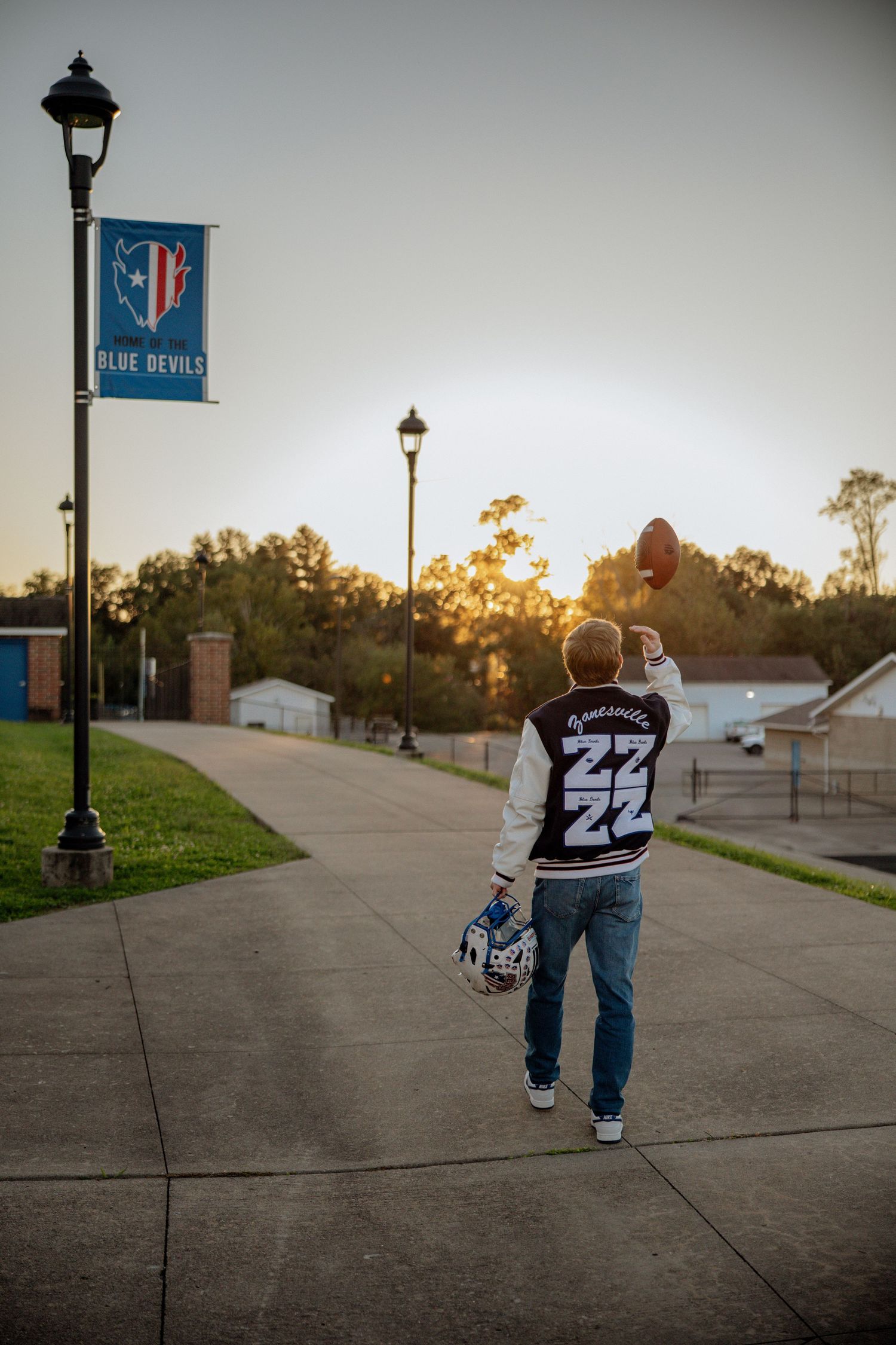 Student in letterman jacket #22 tosses football at sunset near Blue Devils banner on school campus pathway.