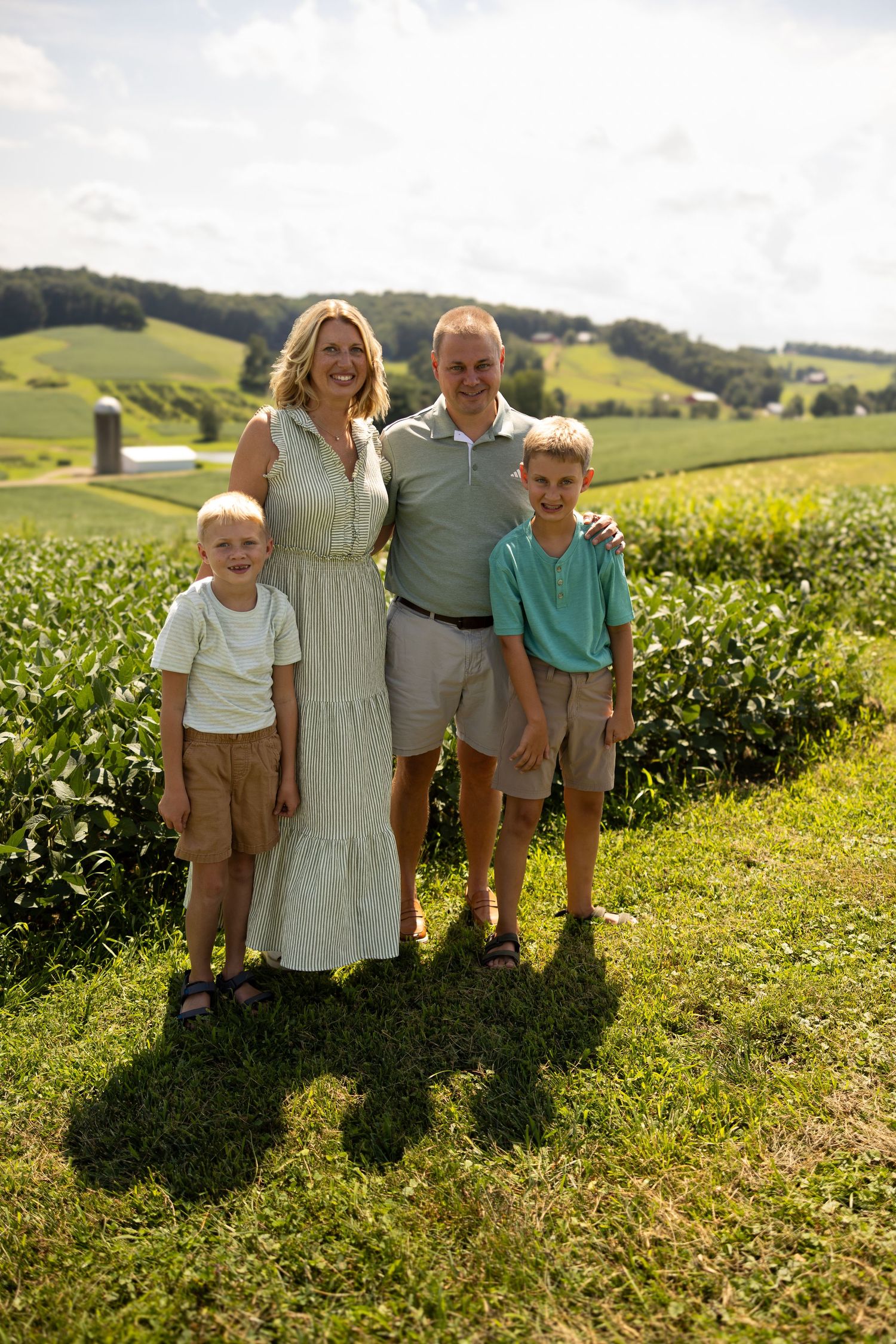 A happy family of four poses outdoors on a sunny day with lush green farmland and a red barn in the background.