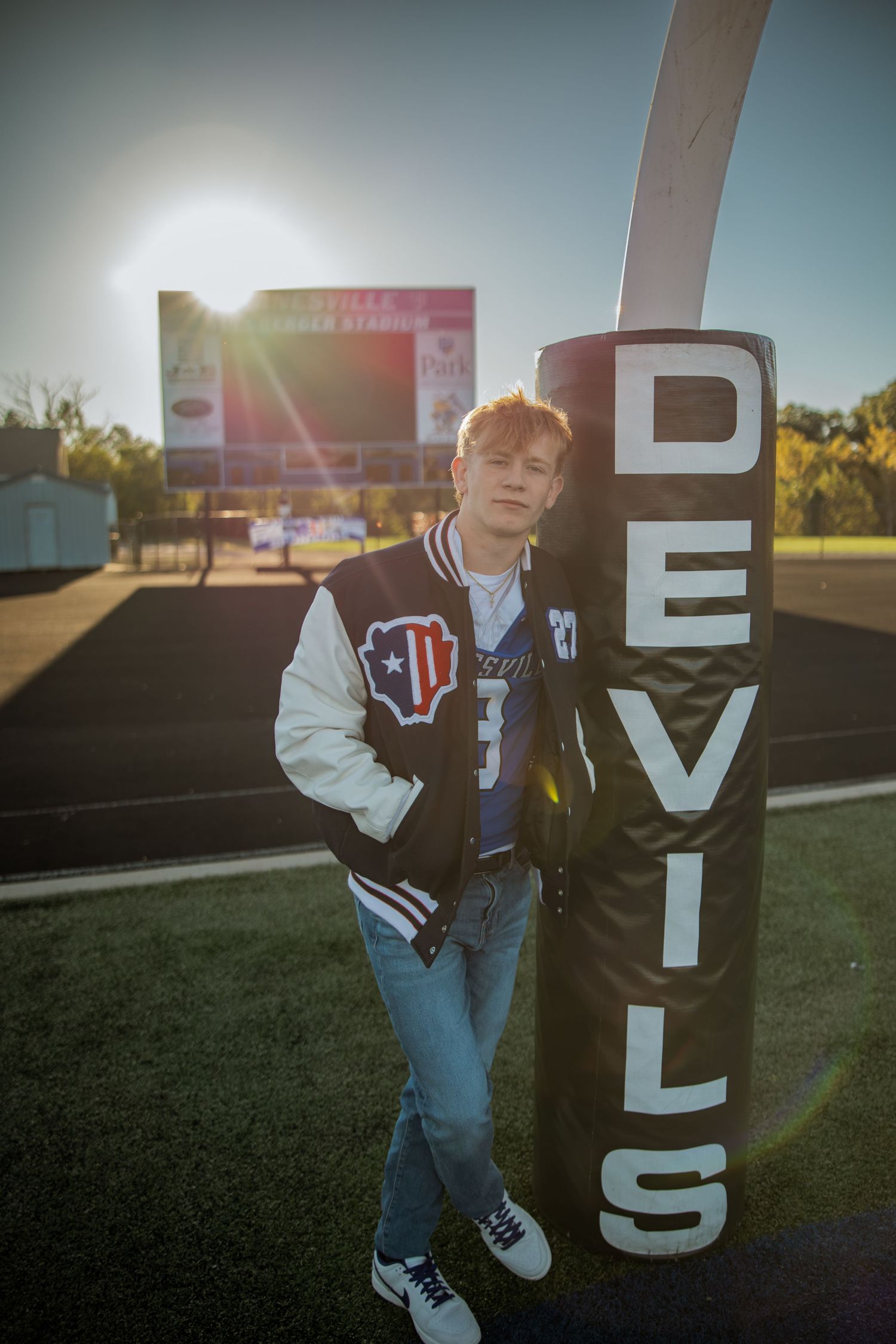 Teen in varsity jacket leans against Devils football field post at sunset on a high school athletic field.