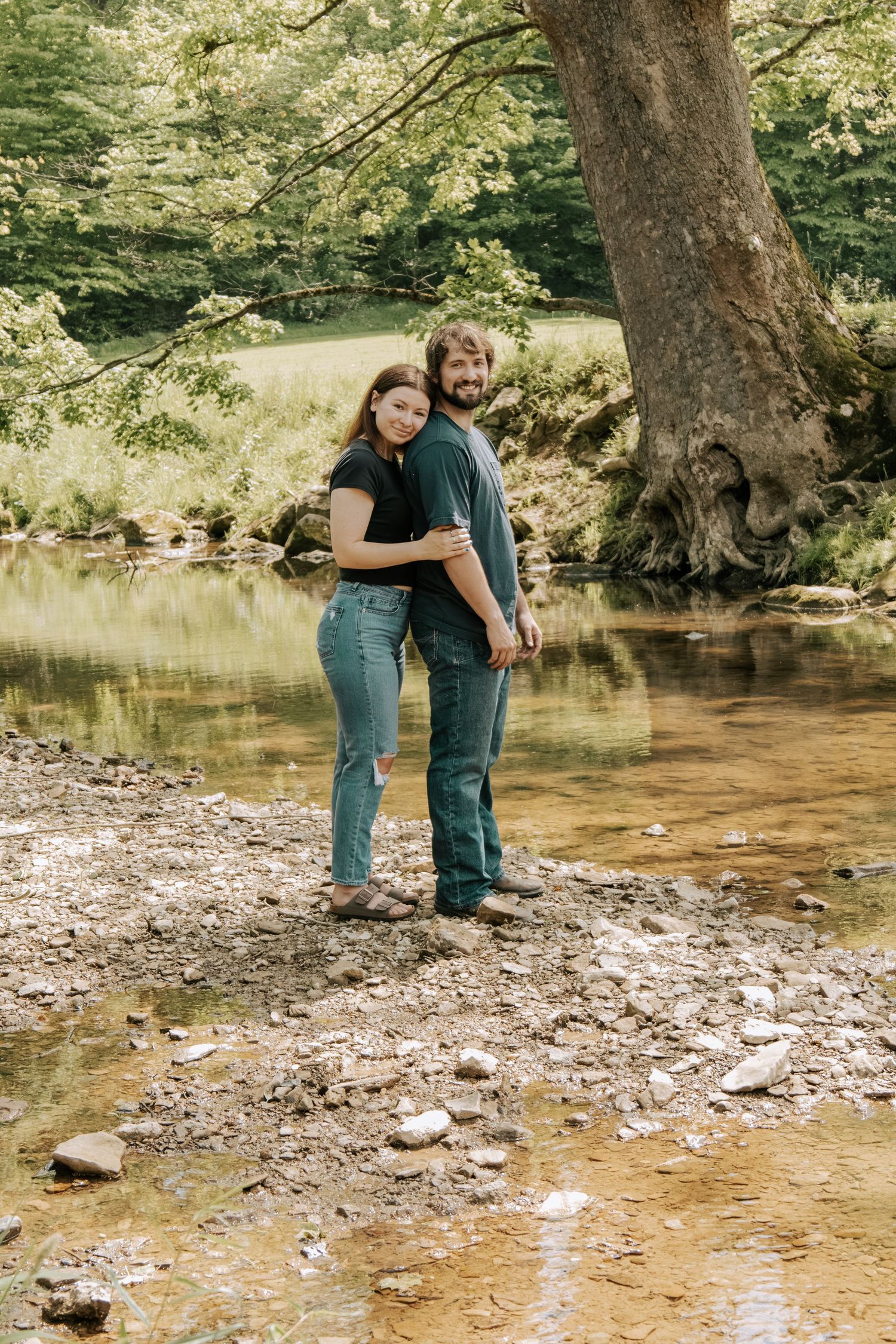 Couple embracing by a rocky creek under a large tree in a lush green forest setting.