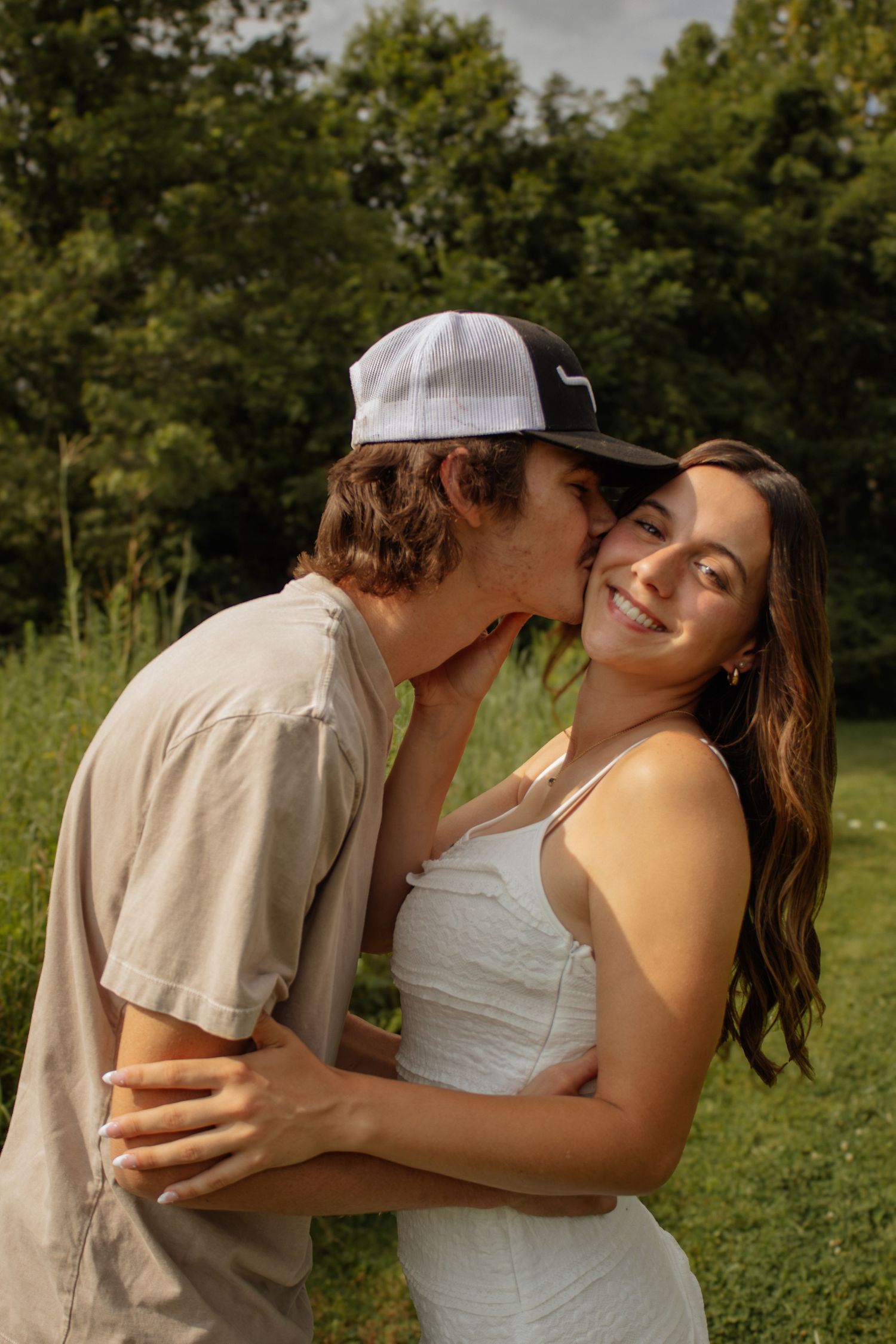 Young couple sharing a romantic moment outdoors, man kissing woman's cheek while she smiles, green trees background.