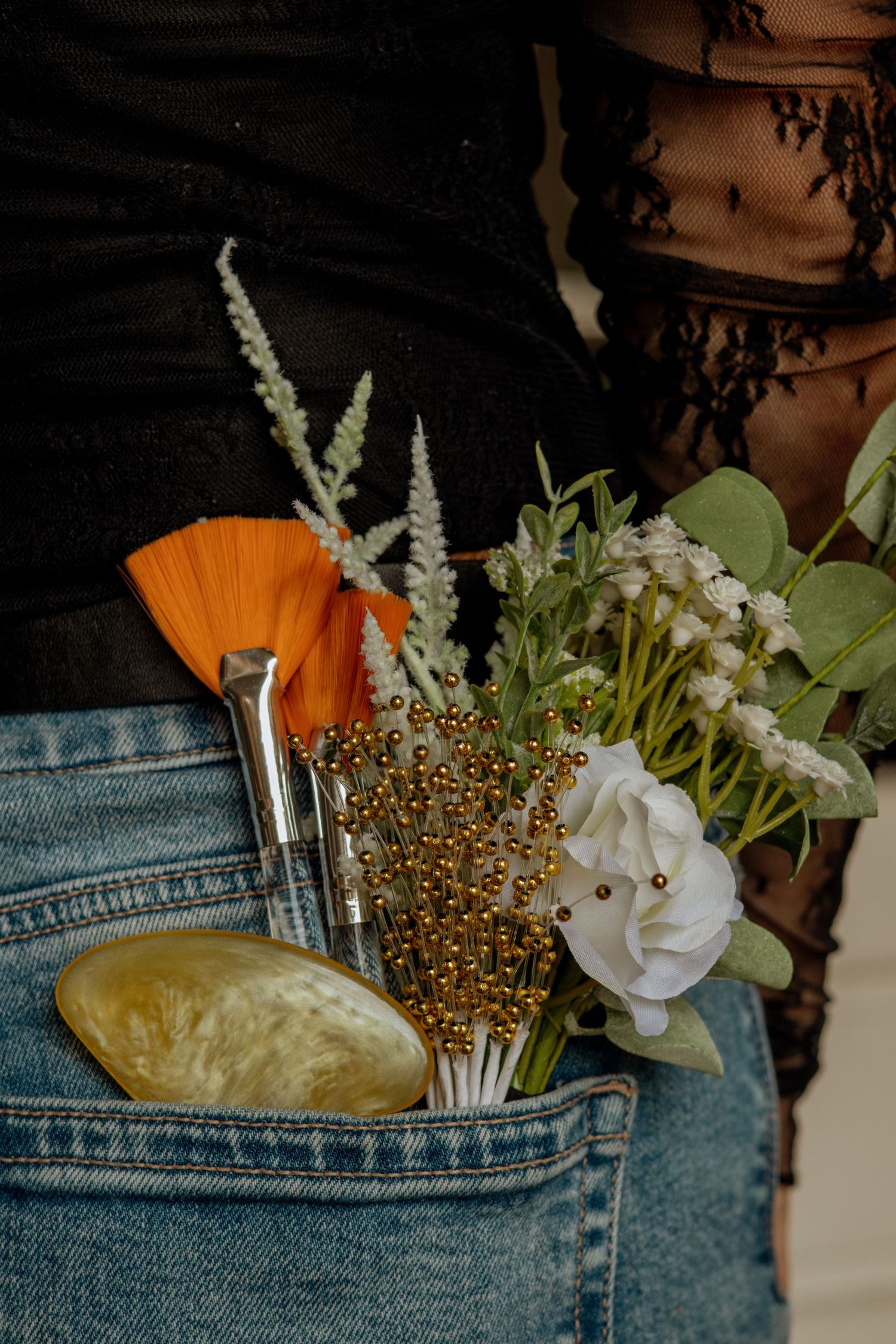 Beauty tools, flowers, and gold beads tucked into back pocket of denim jeans worn by woman.