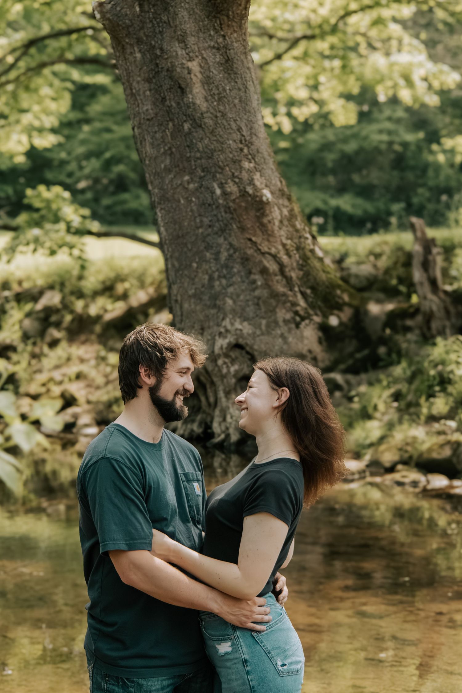 Smiling couple embracing near a creek surrounded by lush green trees in a romantic outdoor portrait.