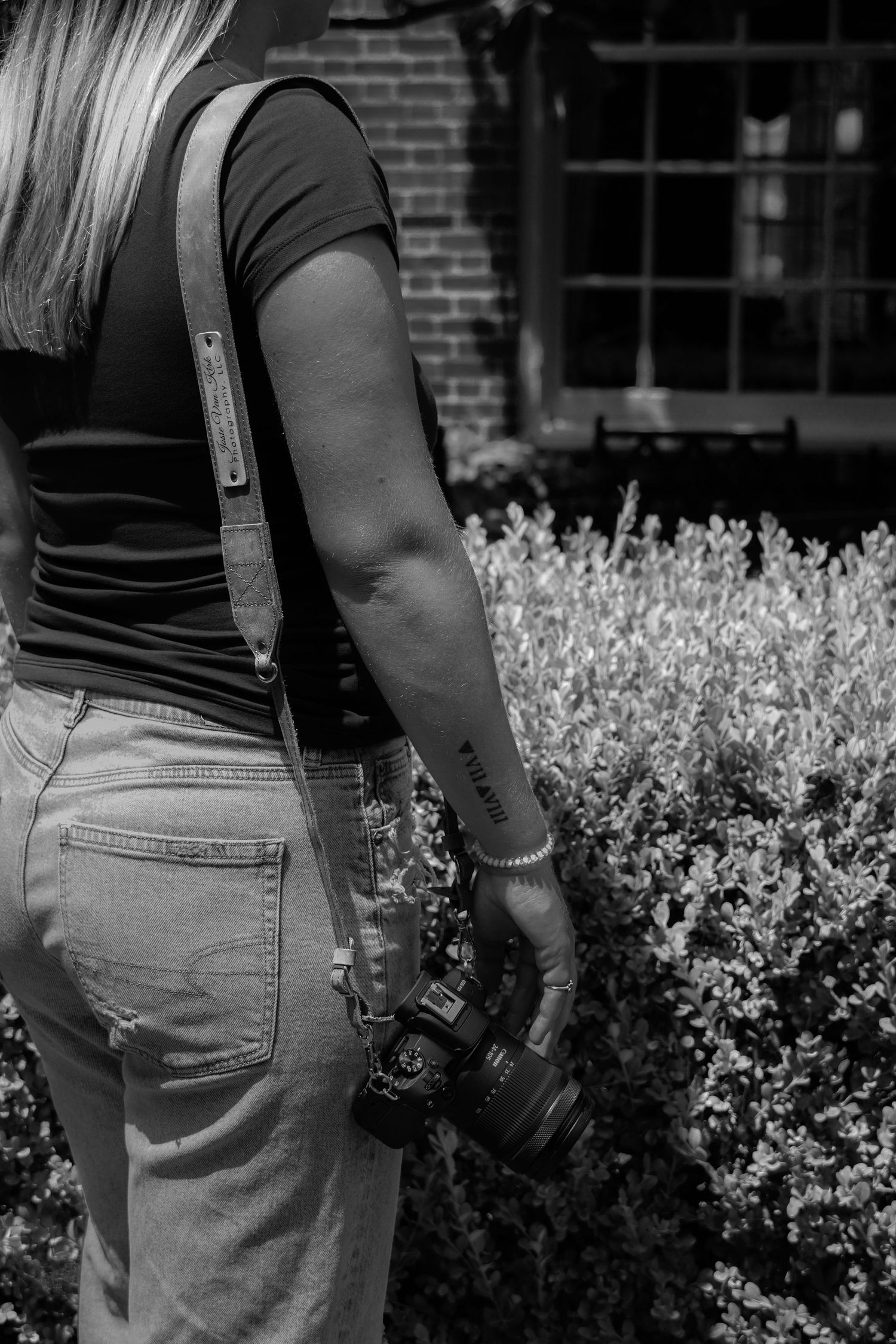 Black and white photo of female photographer holding Canon camera with leather strap outdoors.