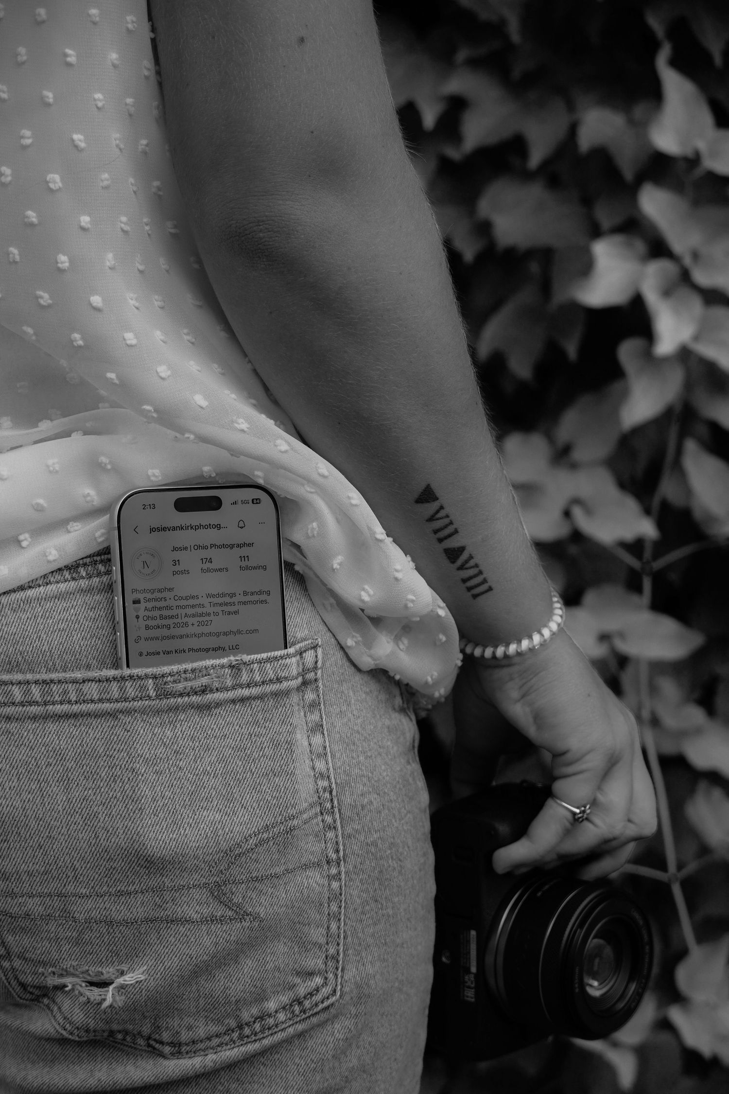 Black and white photo of woman with Roman numeral wrist tattoo, pearl bracelet, holding camera, phone in jeans pocket.