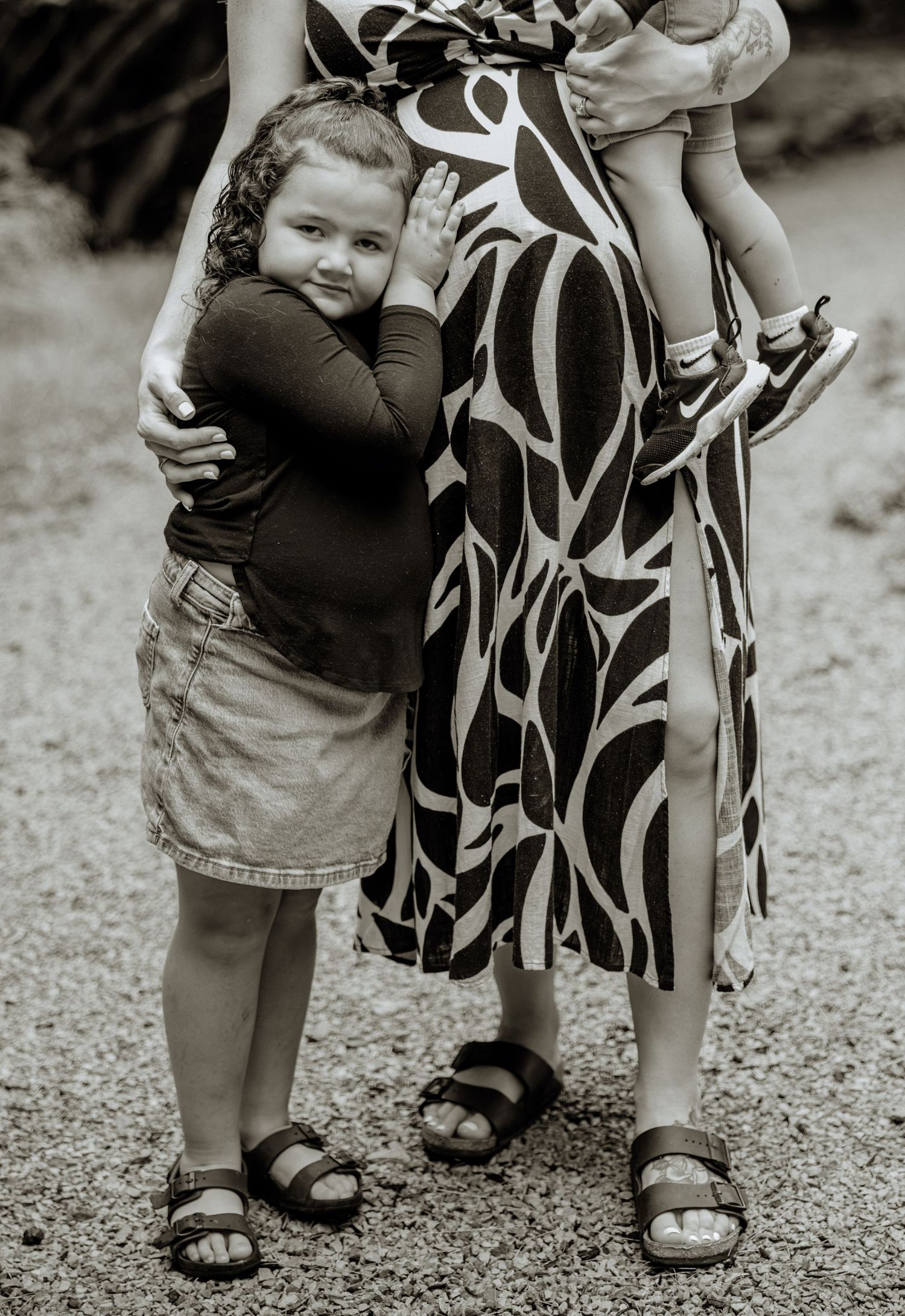 Black and white photo of a young girl hugging her mother in a patterned dress while a toddler is held on her hip.
