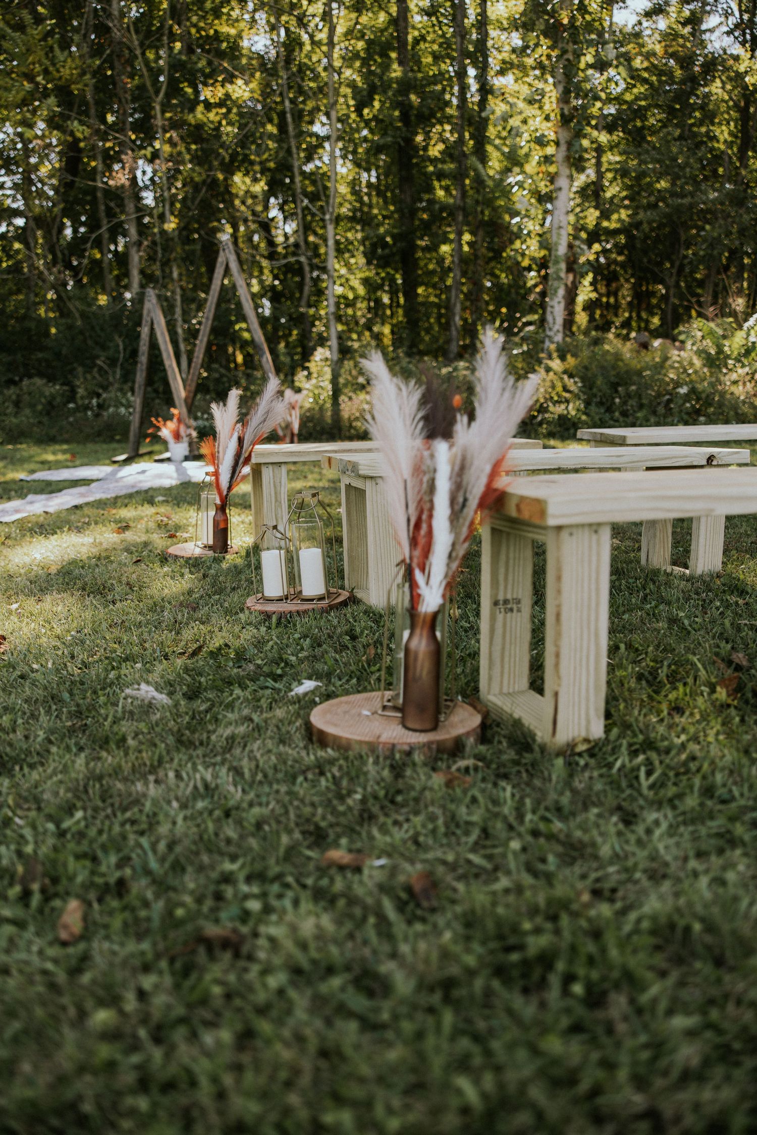 Rustic outdoor wedding ceremony aisle with wooden benches, pampas grass in brown bottles, and lanterns on wood slices.