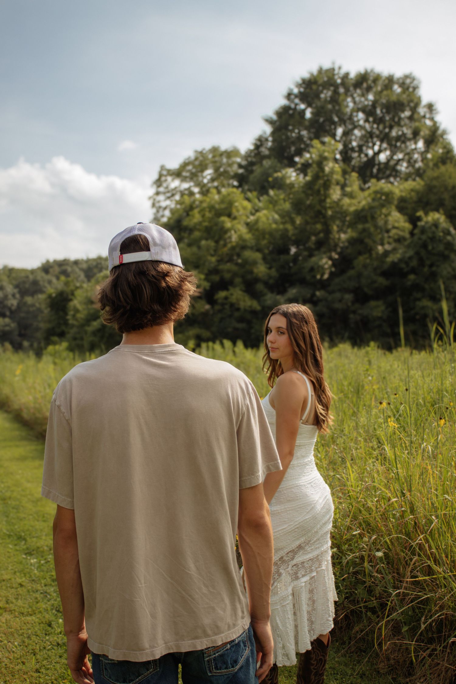 A young couple walks through a lush green field, the woman in a white dress glancing back over her shoulder.