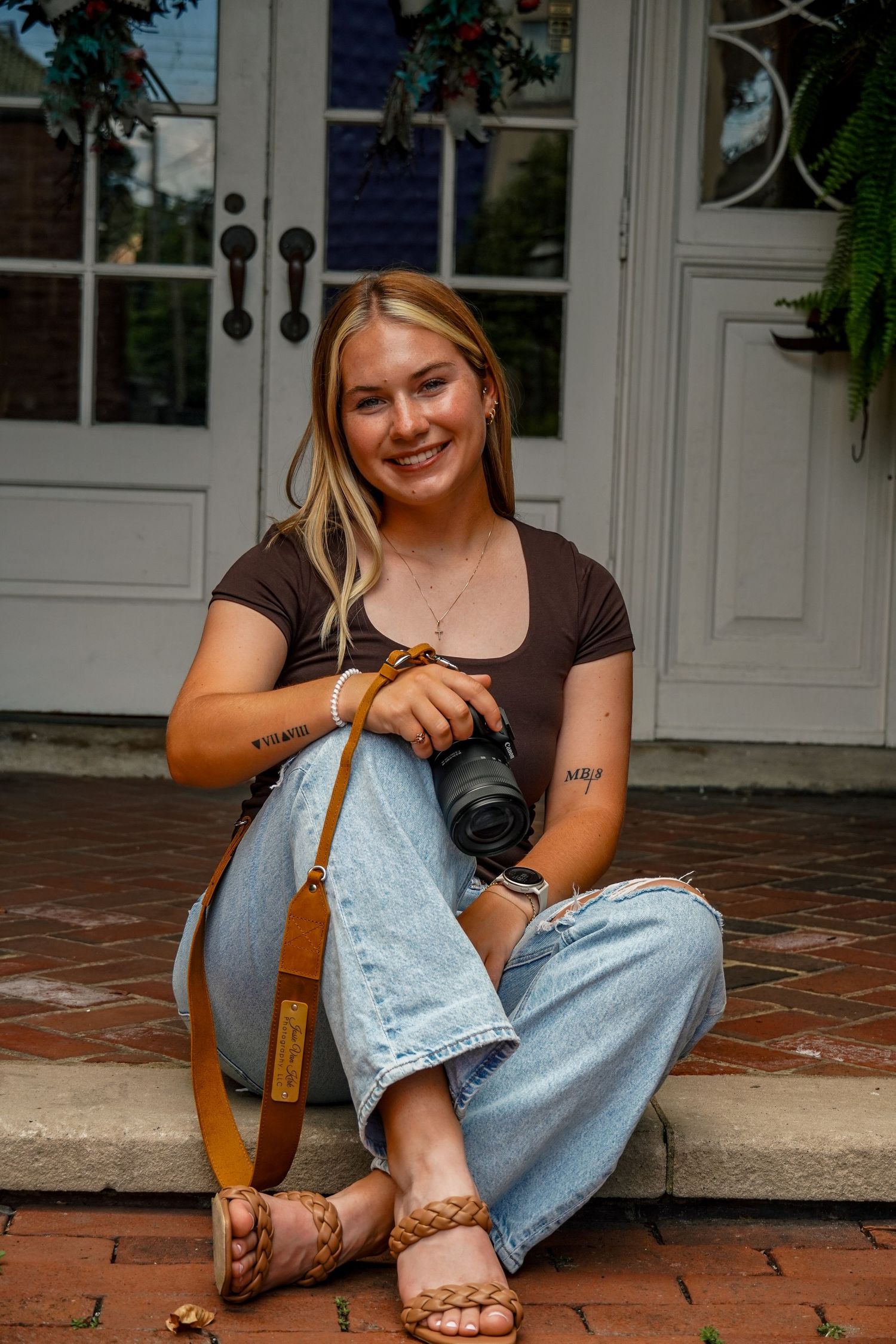 Smiling blonde female photographer sitting on brick steps holding Canon camera with tan leather strap outdoors.
