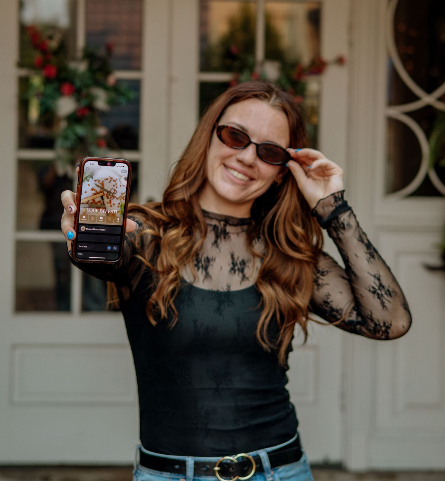 Smiling redhead woman in black lace top holding phone displaying photo while adjusting sunglasses outdoors.
