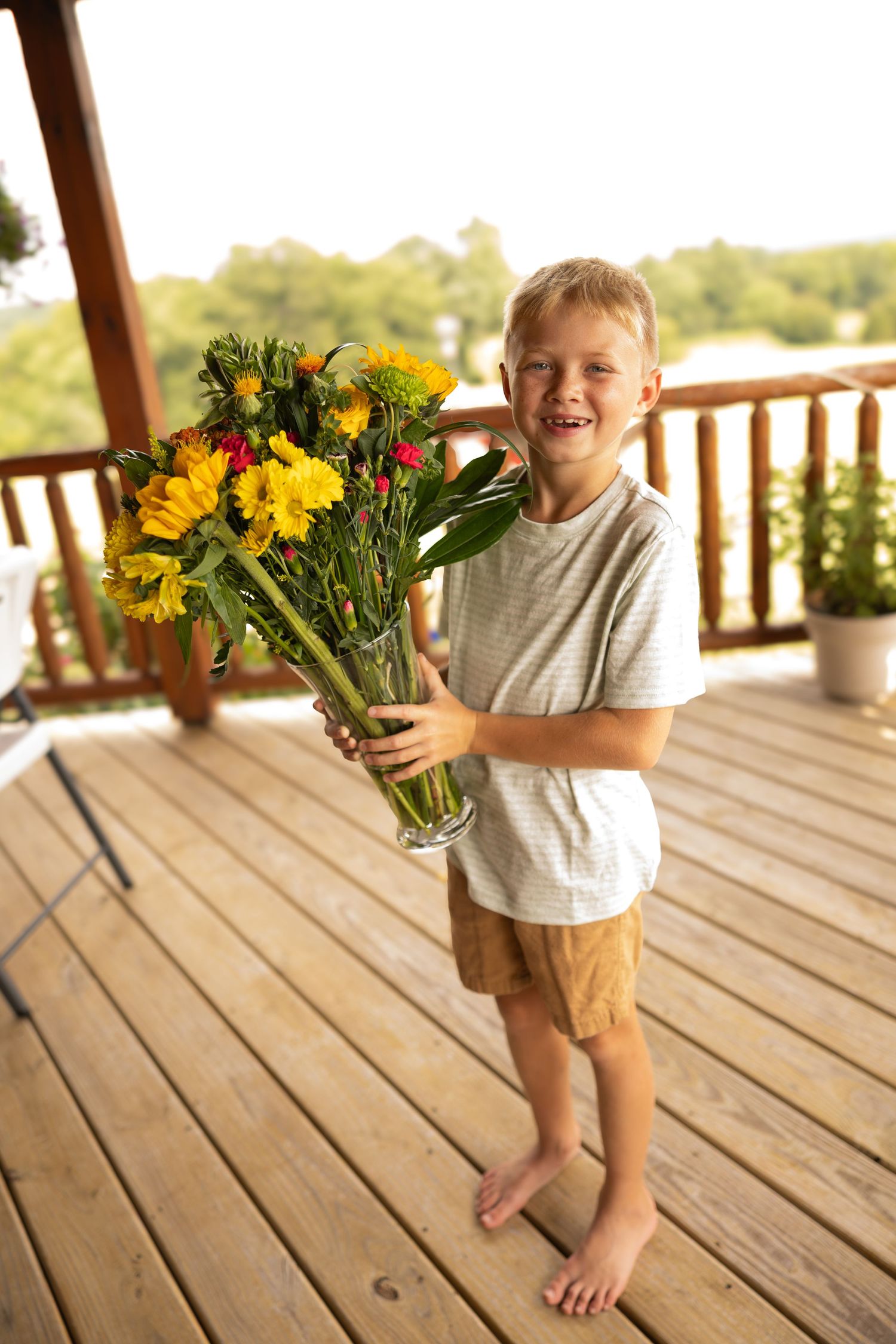 Smiling barefoot boy holding a glass vase of yellow flowers on a wooden deck outdoors.