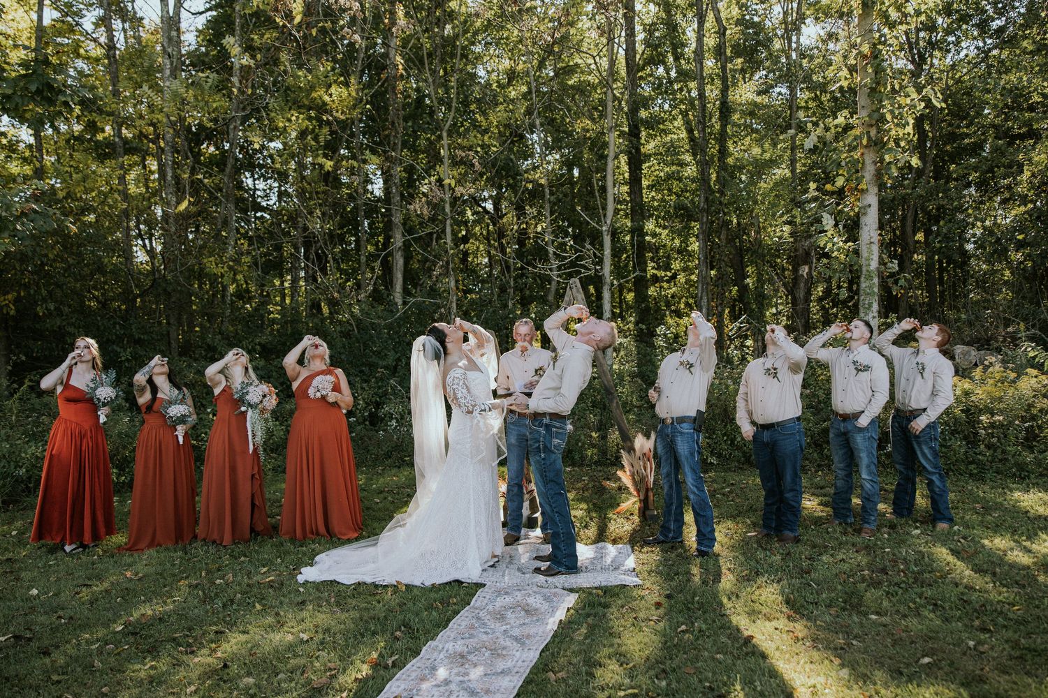 Wedding party in rustic outdoor ceremony, bridesmaids in red dresses, groomsmen in jeans, couple kissing at altar.