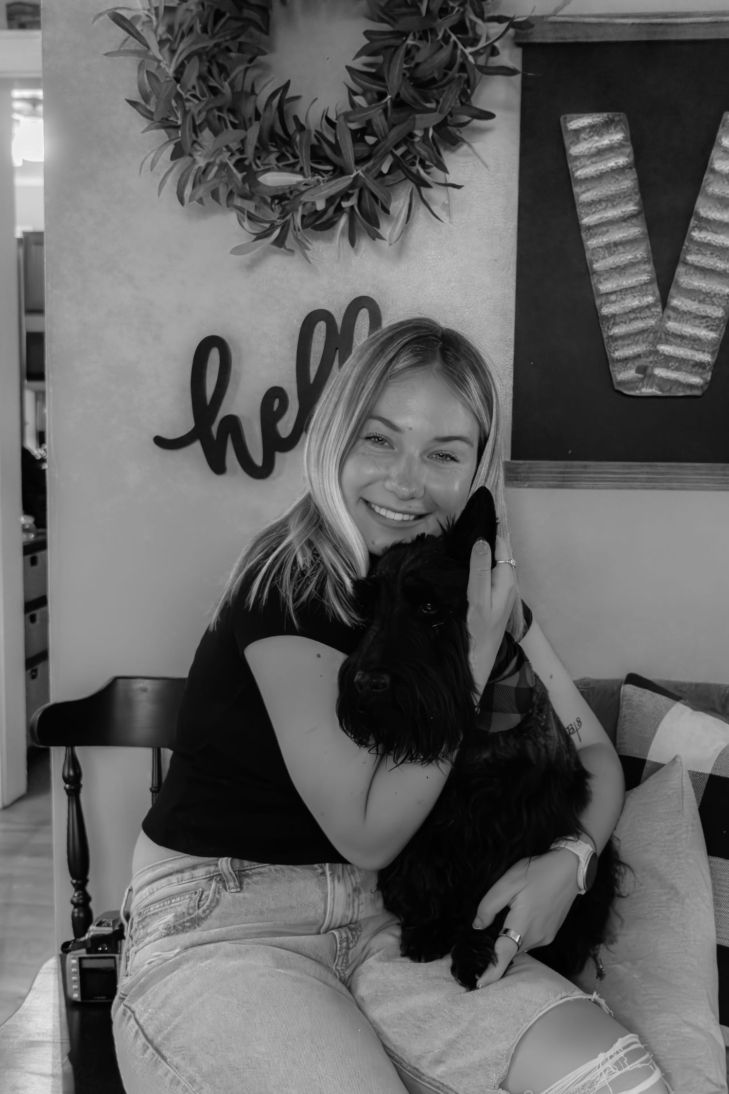 Smiling blonde woman hugging a black dog indoors in black and white portrait photo.