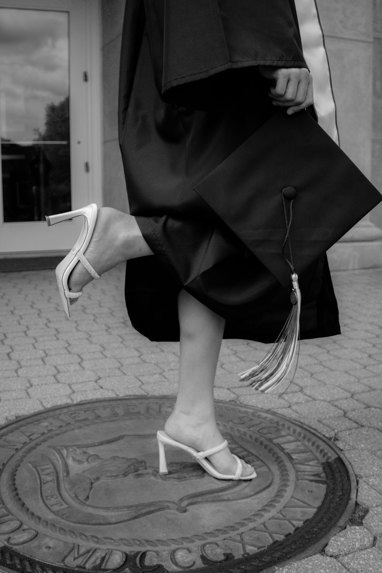 Black and white photo of graduate in gown holding cap while standing on university seal.