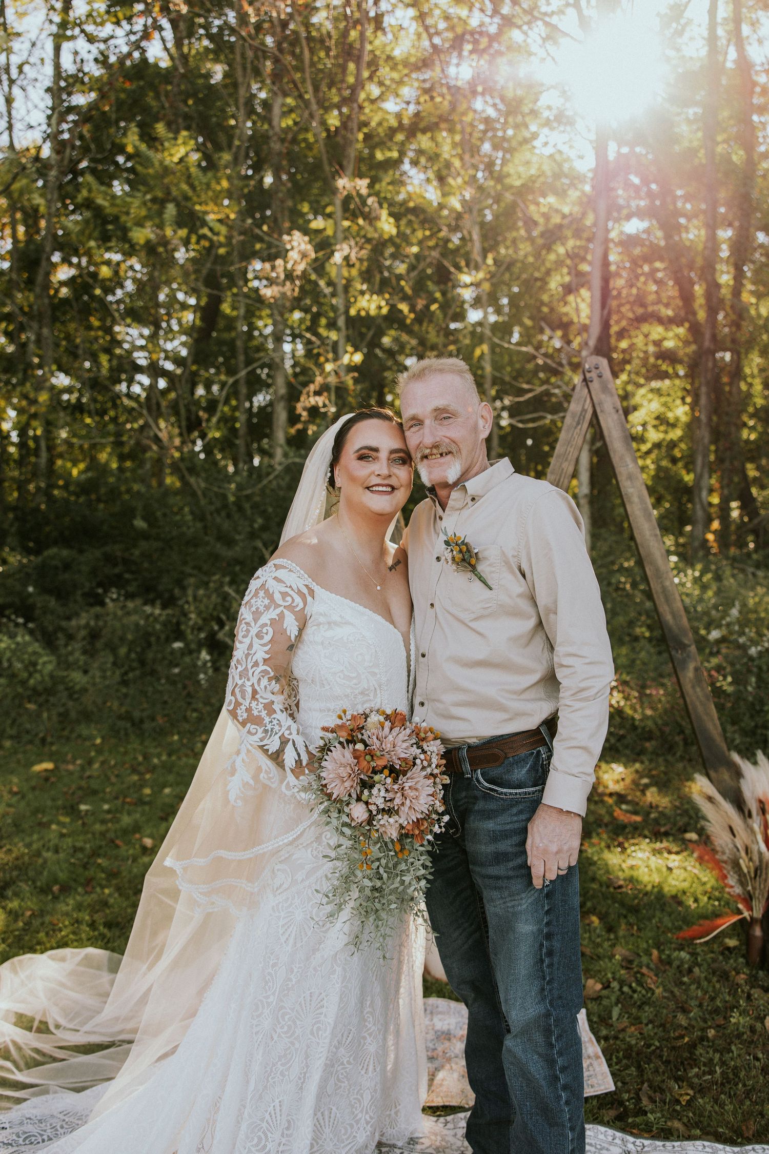 Bride in lace wedding gown holding bouquet poses with older man in jeans and boutonniere at outdoor woodland wedding ceremony.