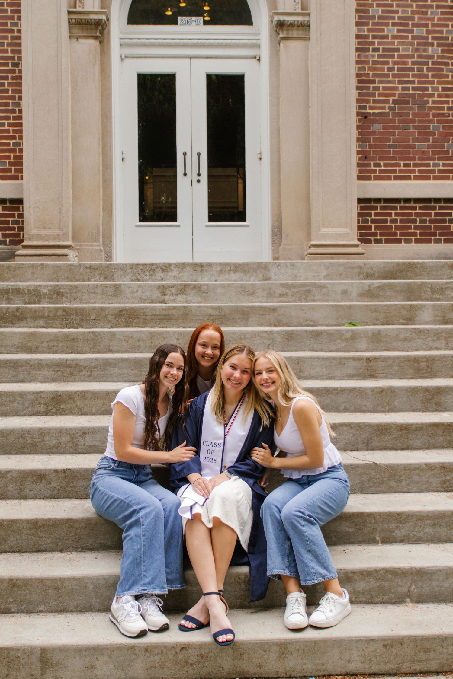 Four young women sitting on college building steps, one in graduation attire.