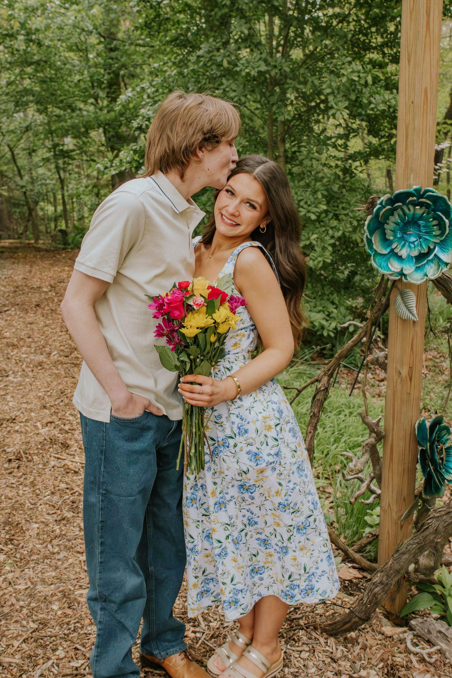 Young couple sharing a kiss outdoors, woman holding colorful bouquet near a decorative garden arch.