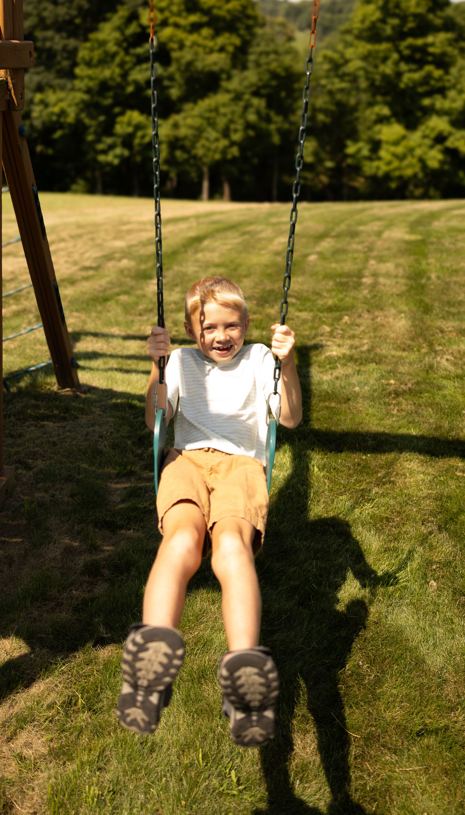 Happy child swinging on a backyard swing set, wearing white shirt and tan shorts, laughing with joy on a sunny day.
