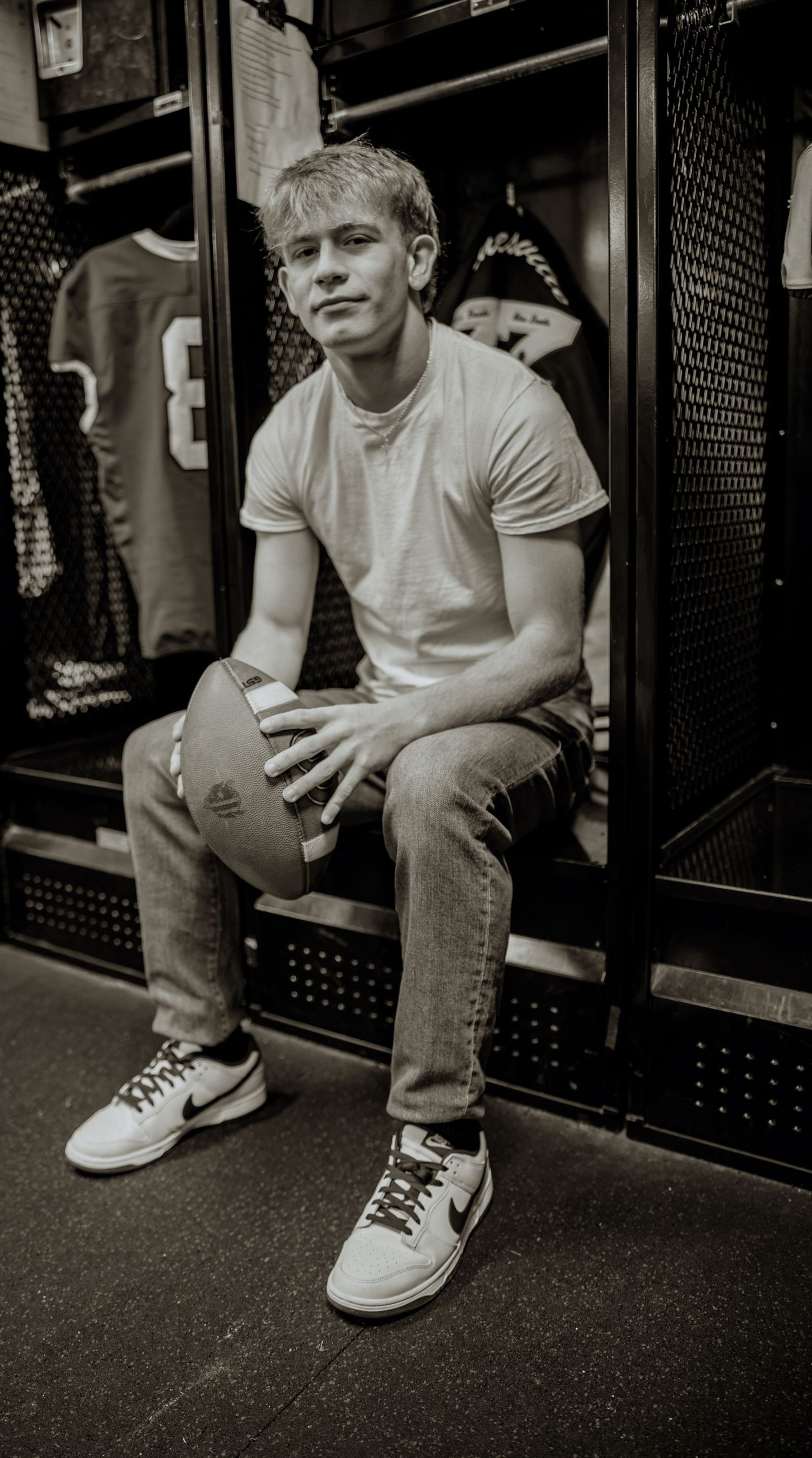 Young male athlete sits in a locker room holding a football, wearing jeans, white tee, and Nike sneakers.